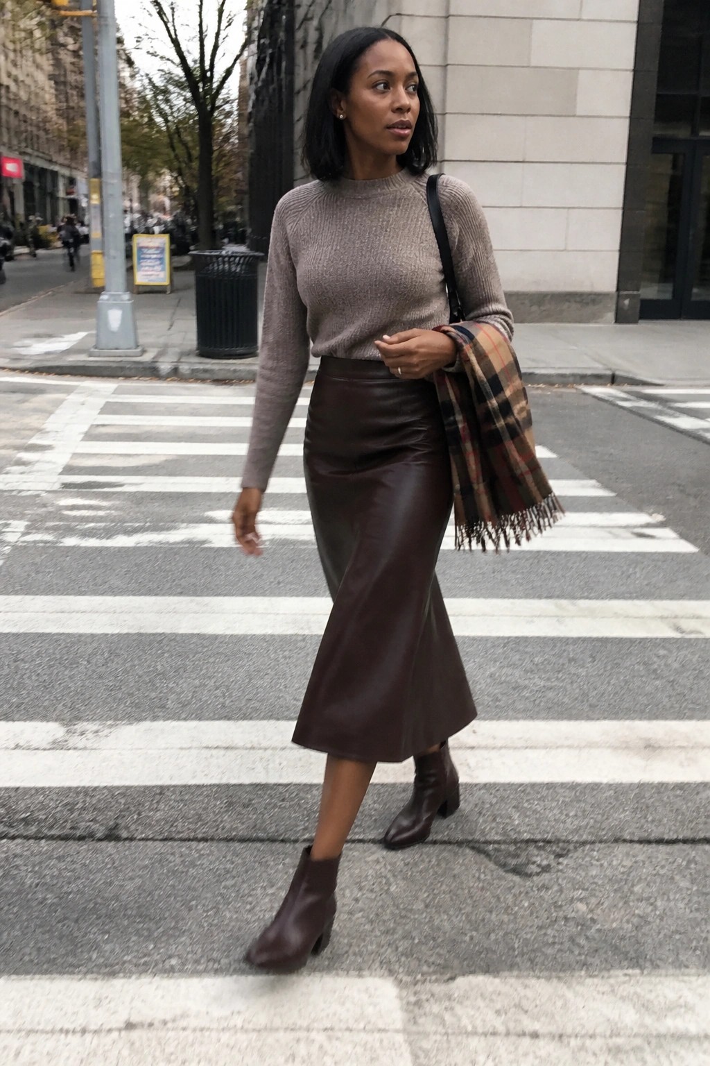 Woman in taupe sweater and brown leather skirt crossing city crosswalk with plaid scarf