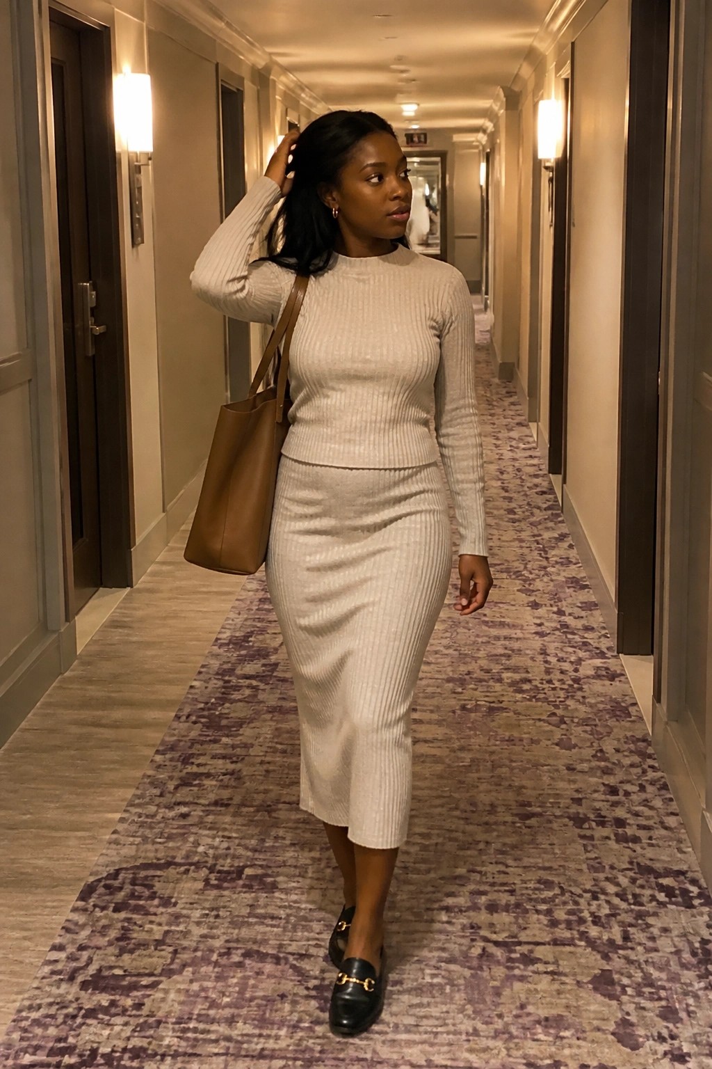 Woman in beige ribbed sweater and skirt with brown tote in hotel hallway