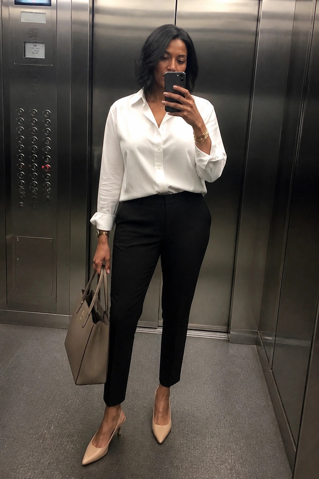 Black woman in elevator mirror selfie wearing white button down shirt tucked into black slim trousers with nude pointed heels and beige tote bag plus gold jewelry.