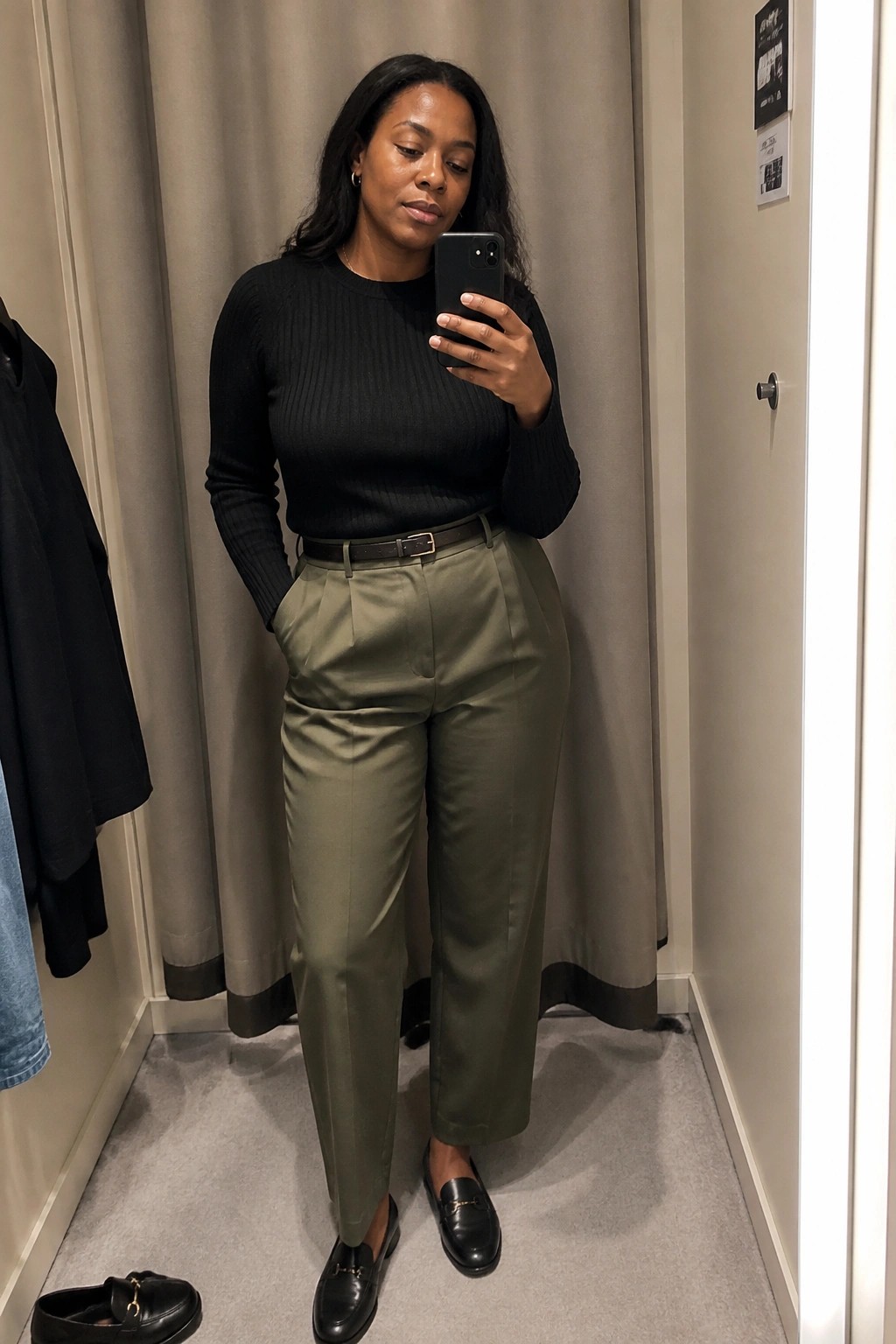 Woman in black sweater and olive pants taking mirror selfie with phone