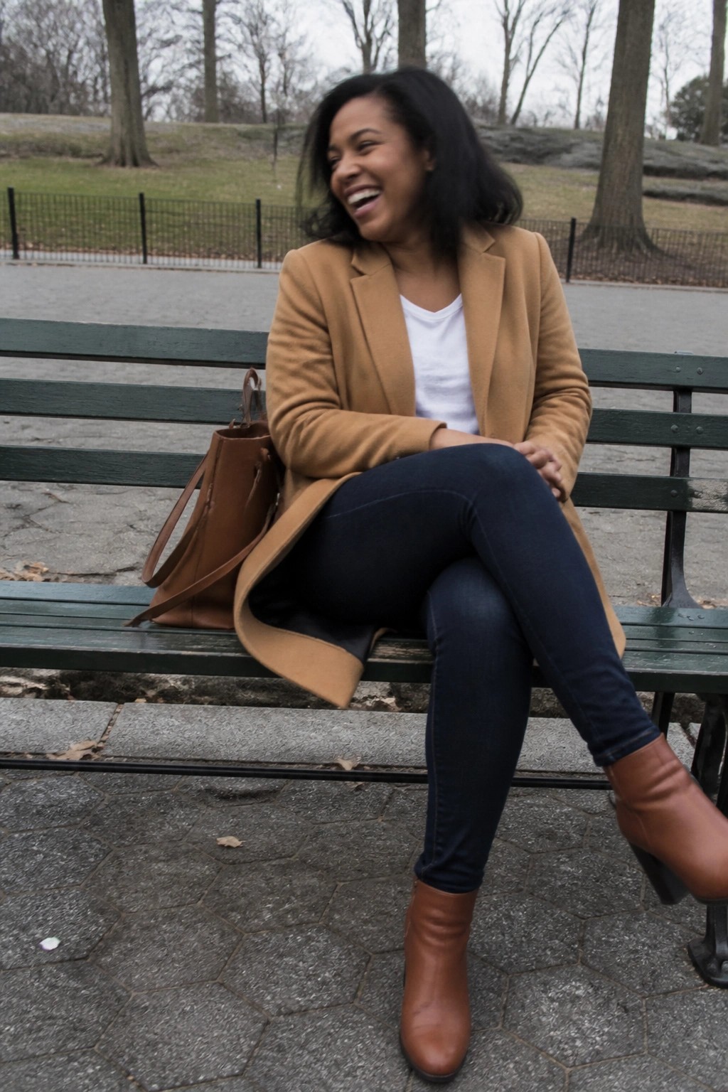 Woman laughing on green park bench in tan coat with brown bag and boots