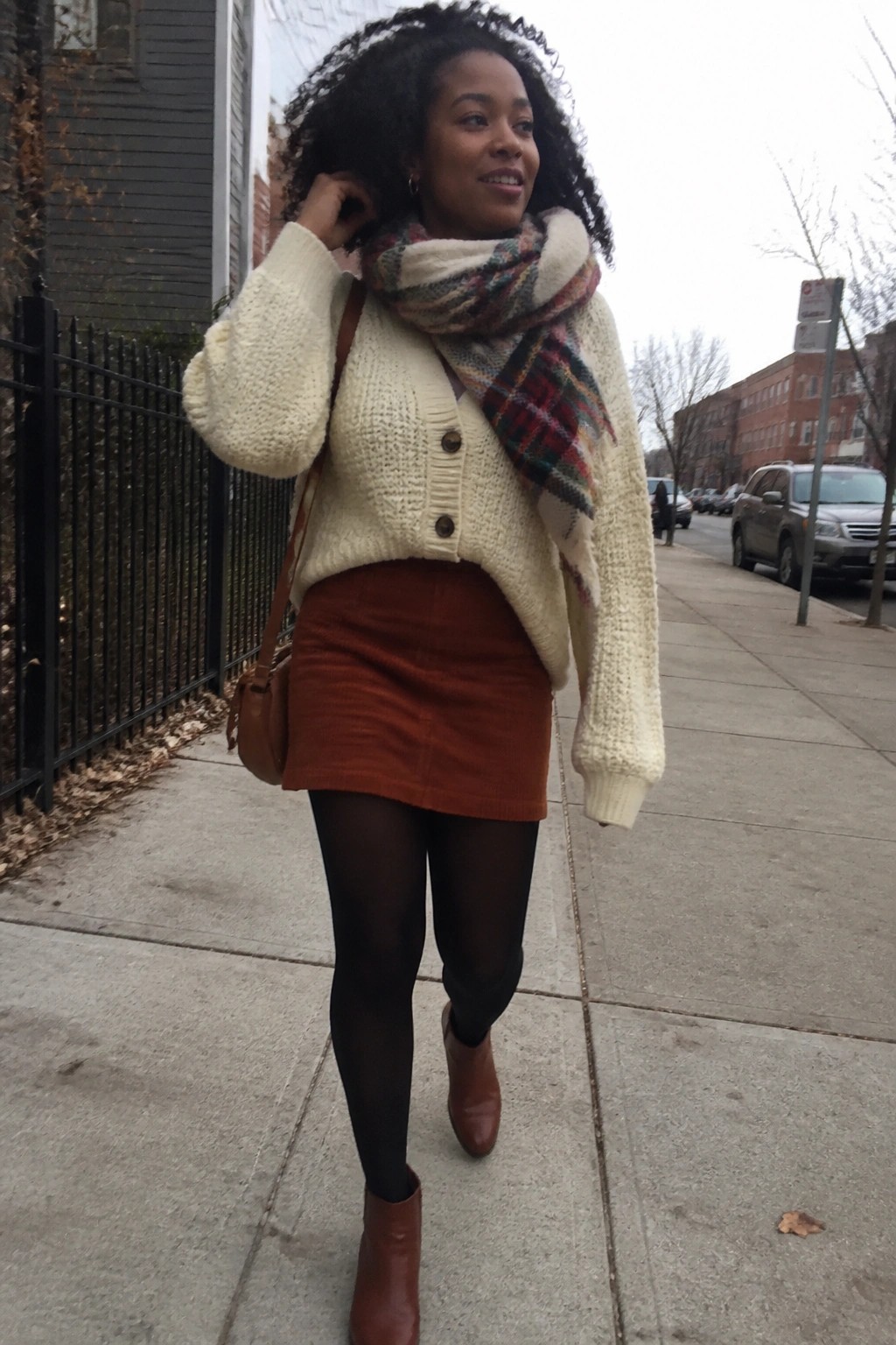 Woman in cream knit cardigan, plaid scarf, rust corduroy skirt and brown boots on sidewalk.