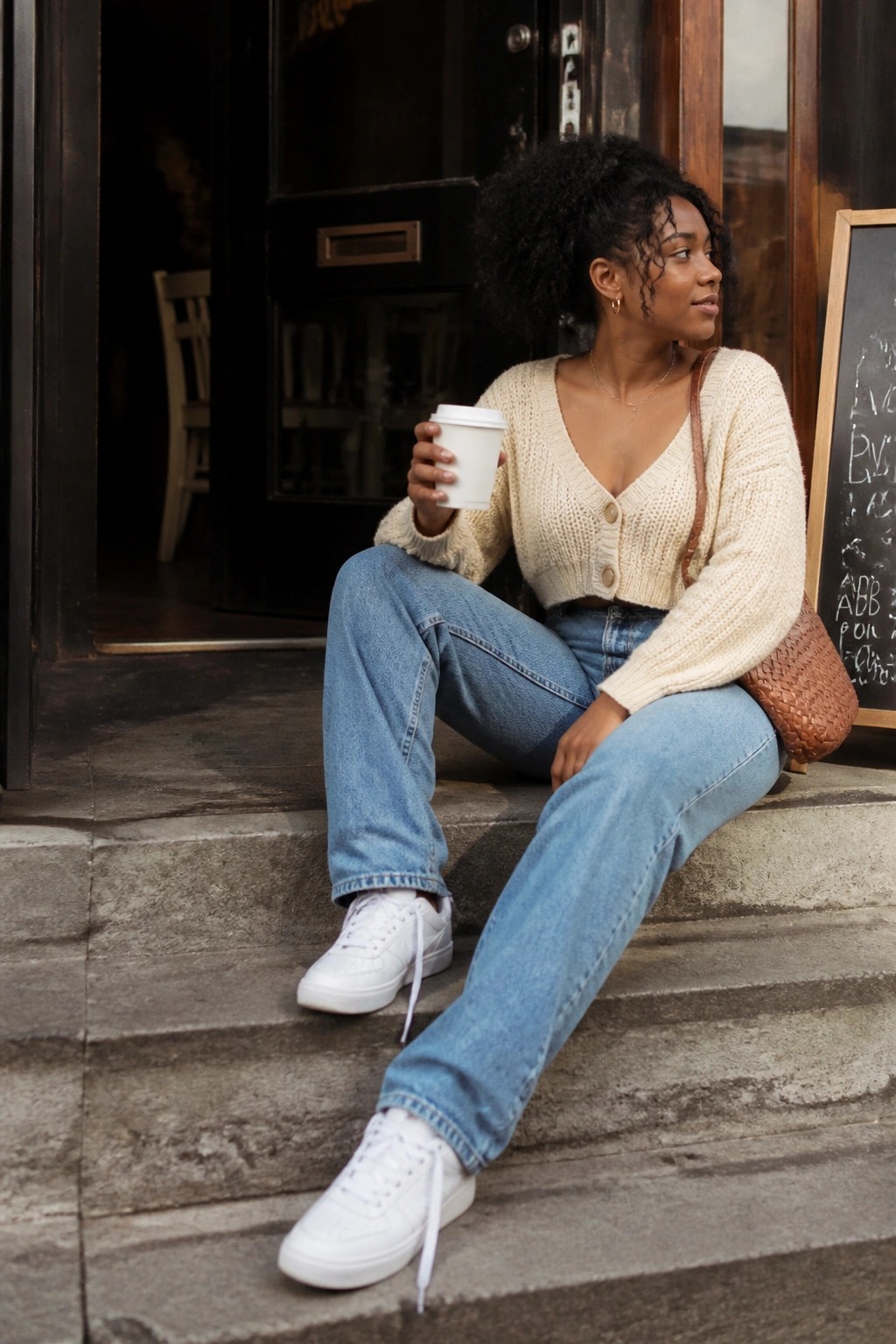 Black woman wearing cream cropped knit cardigan with blue straight leg jeans and white sneakers while holding a coffee cup on stone steps