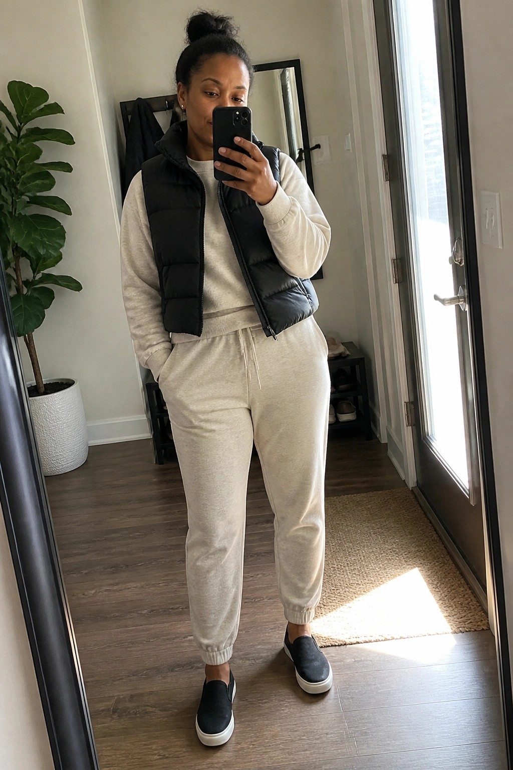 Woman in black puffer vest and beige sweatsuit taking mirror selfie indoors