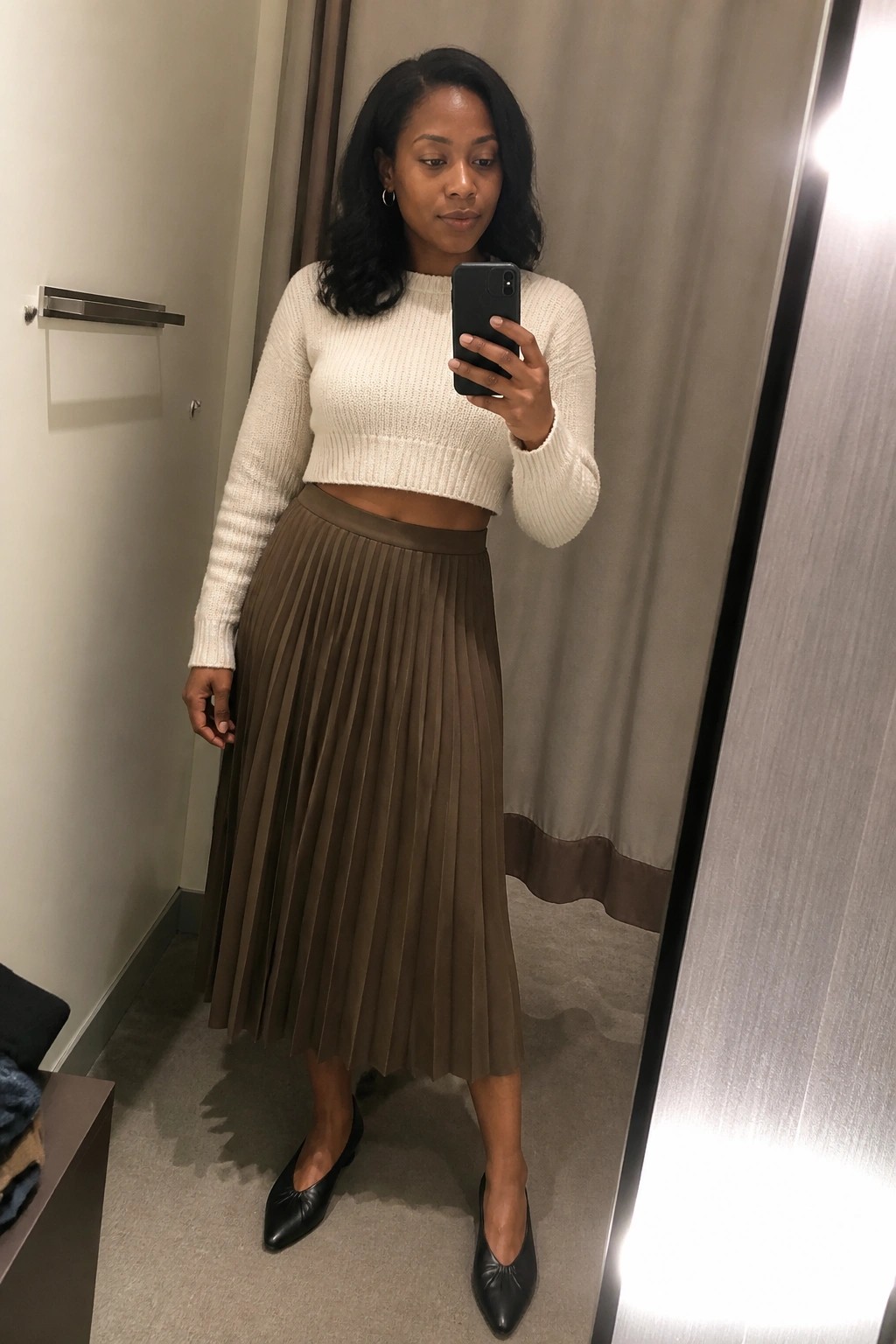 Black woman wearing a white cropped ribbed knit sweater with a taupe pleated midi skirt and black pointed toe flats while taking a mirror selfie.