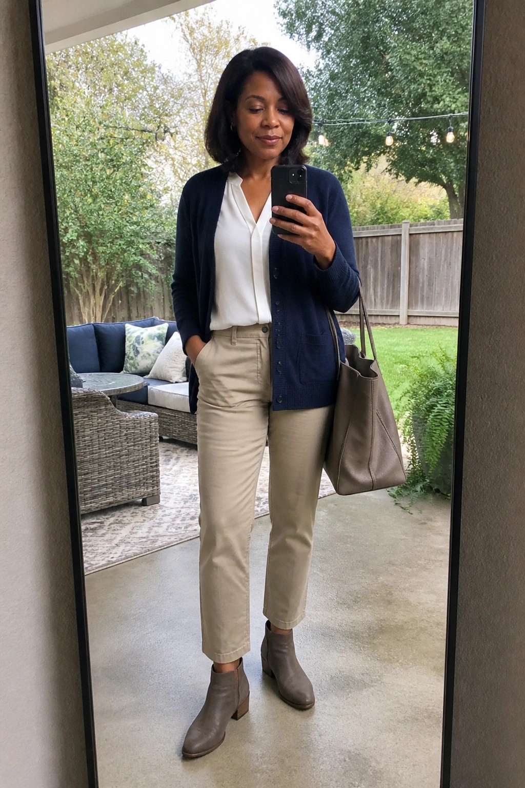 Woman in navy cardigan over white blouse with khaki pants and taupe ankle boots holding a matching tote bag in a mirror selfie.