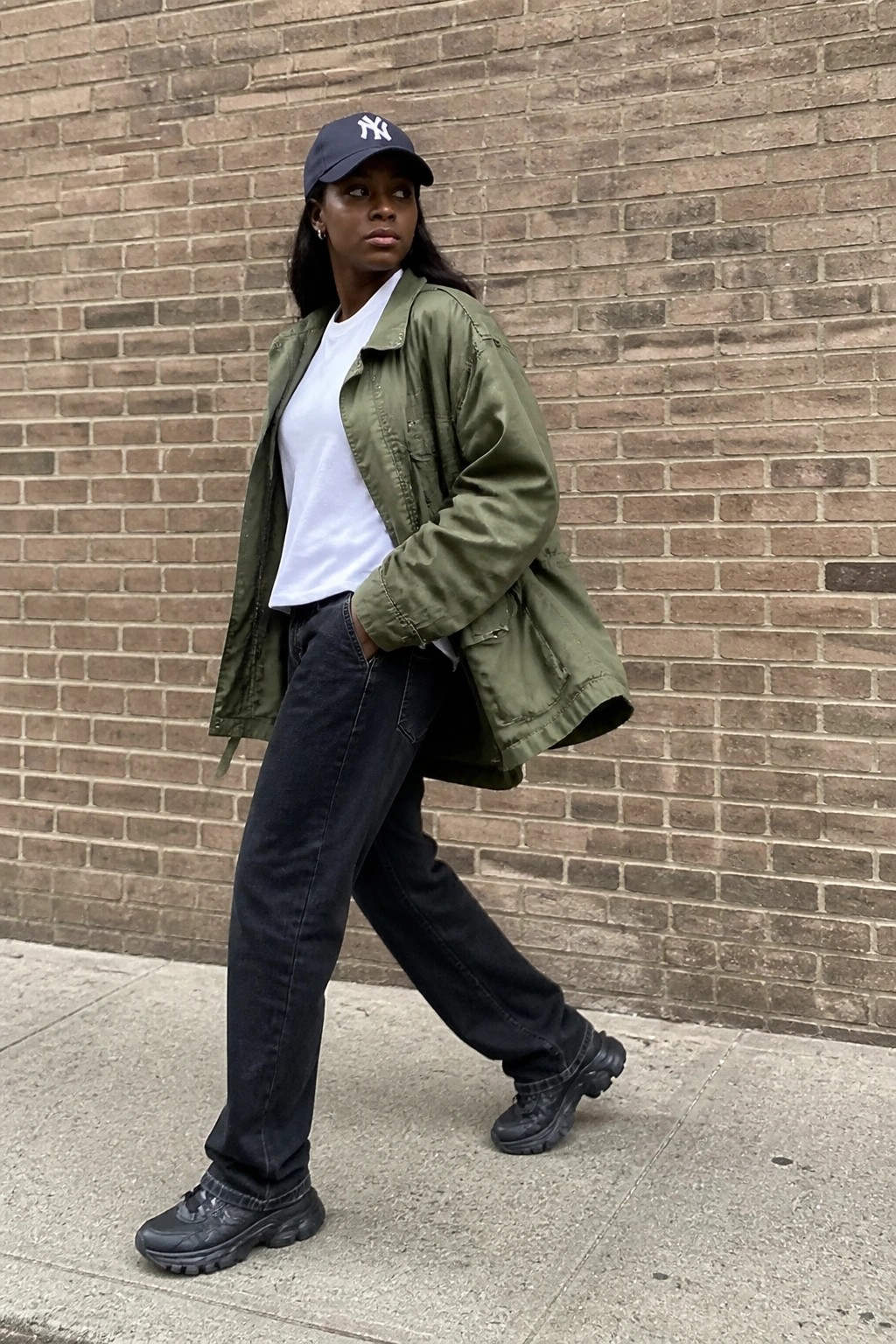 Woman in green jacket, white t-shirt, black jeans, and NY cap by brick wall
