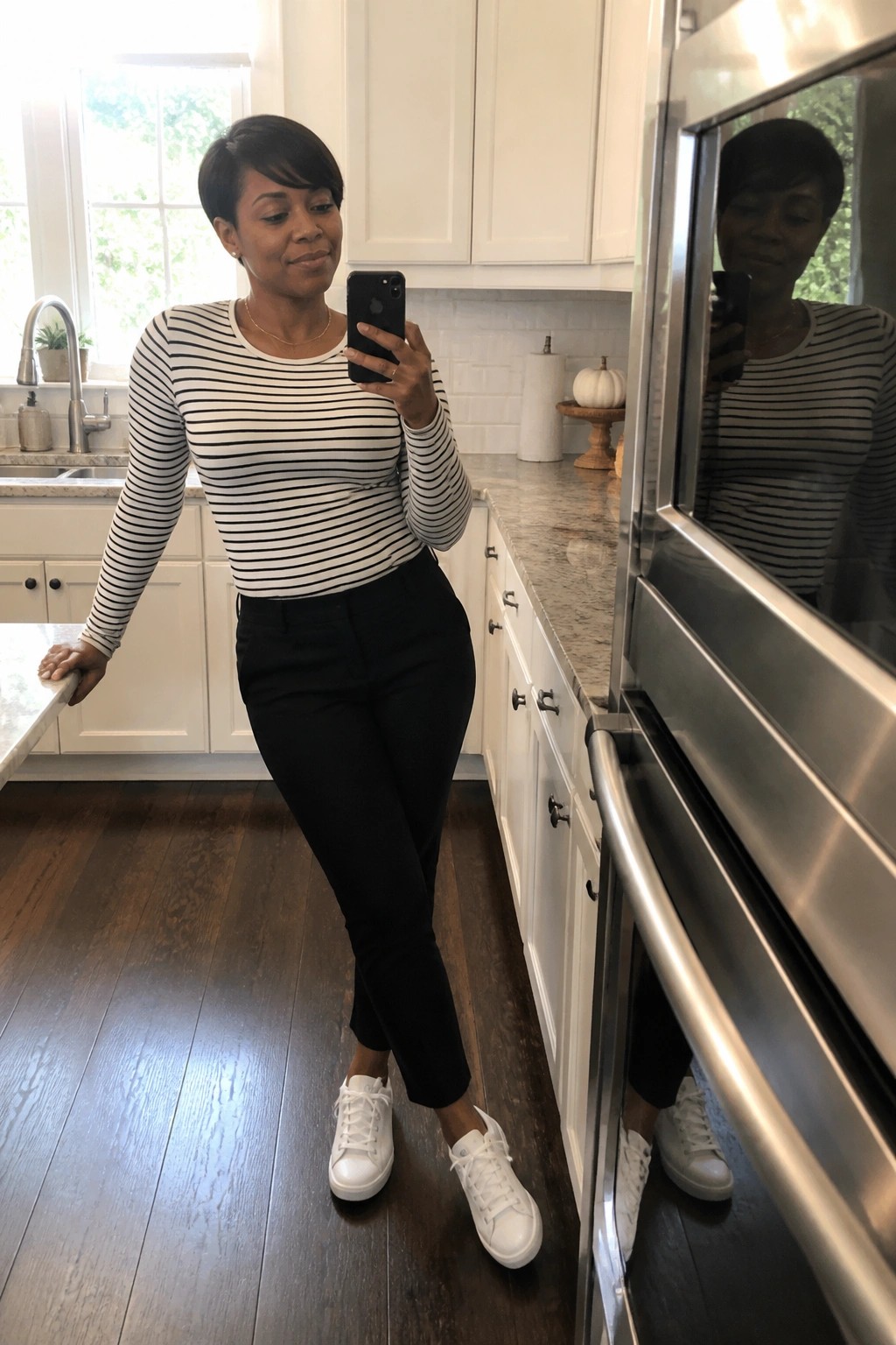 Woman wearing a black and white horizontal striped long sleeve top tucked into black slim pants with white sneakers and a thin gold necklace standing in a kitchen taking a mirror selfie.