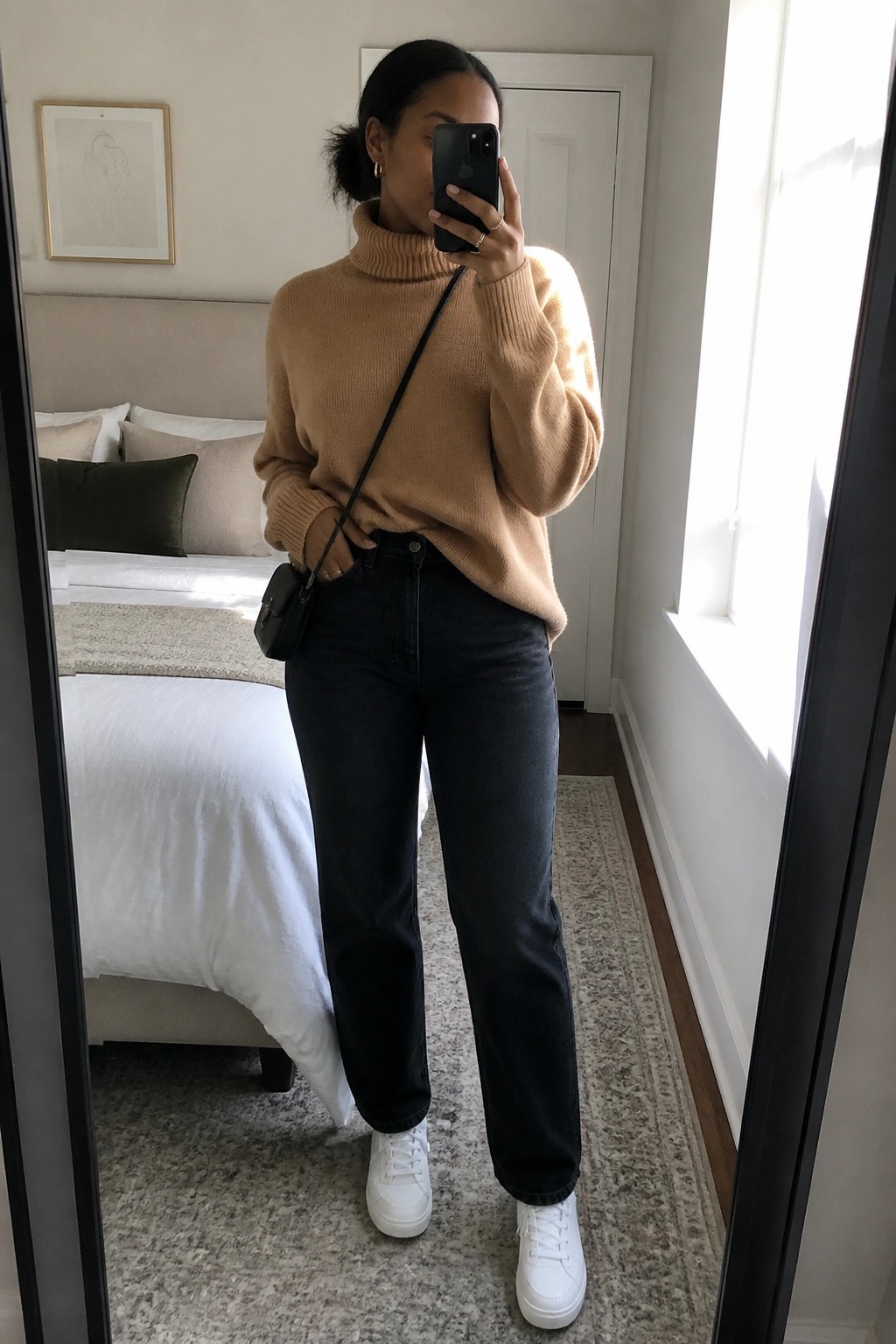 Woman in tan turtleneck sweater black straight leg jeans and white sneakers holding phone for mirror selfie with small black bag