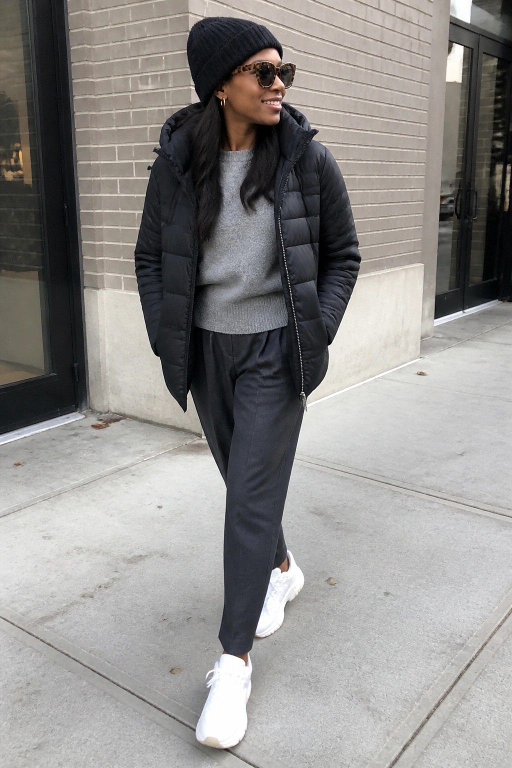 Woman wearing black puffer vest over grey sweater with black pants and white sneakers, black beanie and sunglasses, standing outdoors.