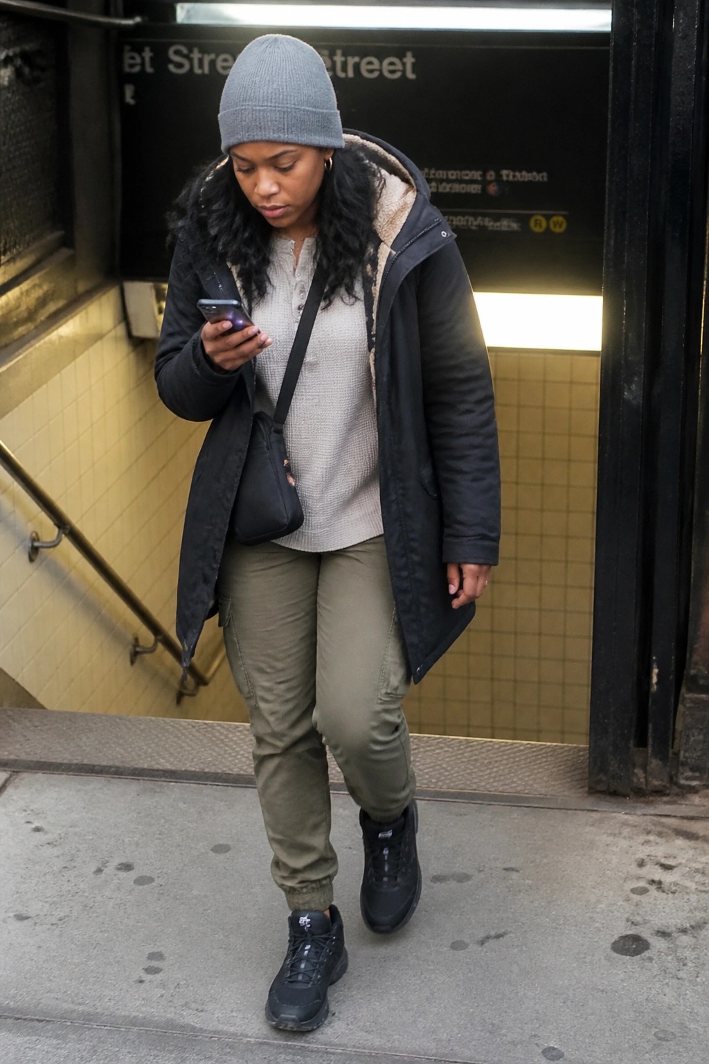 Woman in gray beanie and black coat views smartphone outside subway entrance