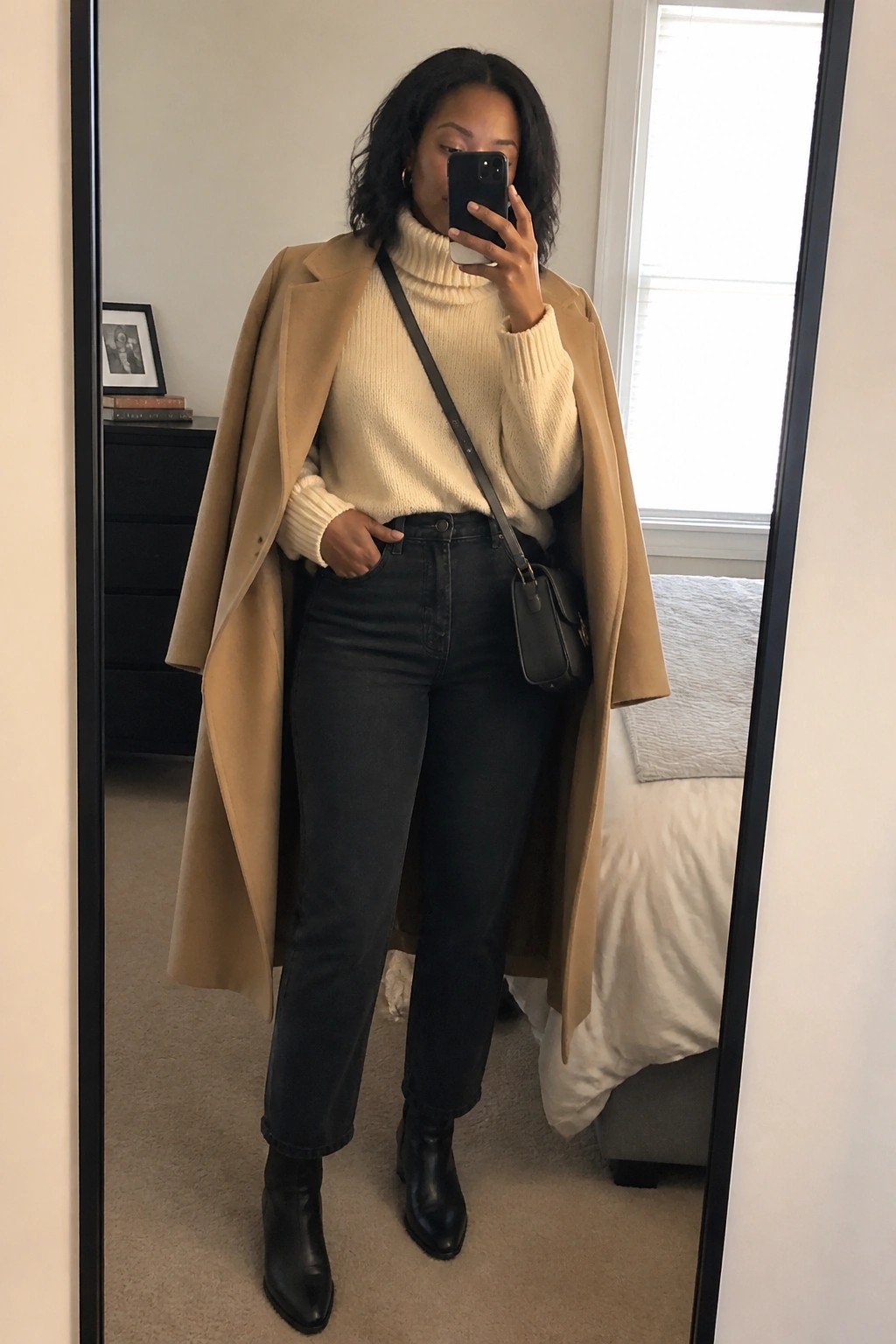 Woman in tan coat and cream turtleneck taking mirror selfie in bedroom