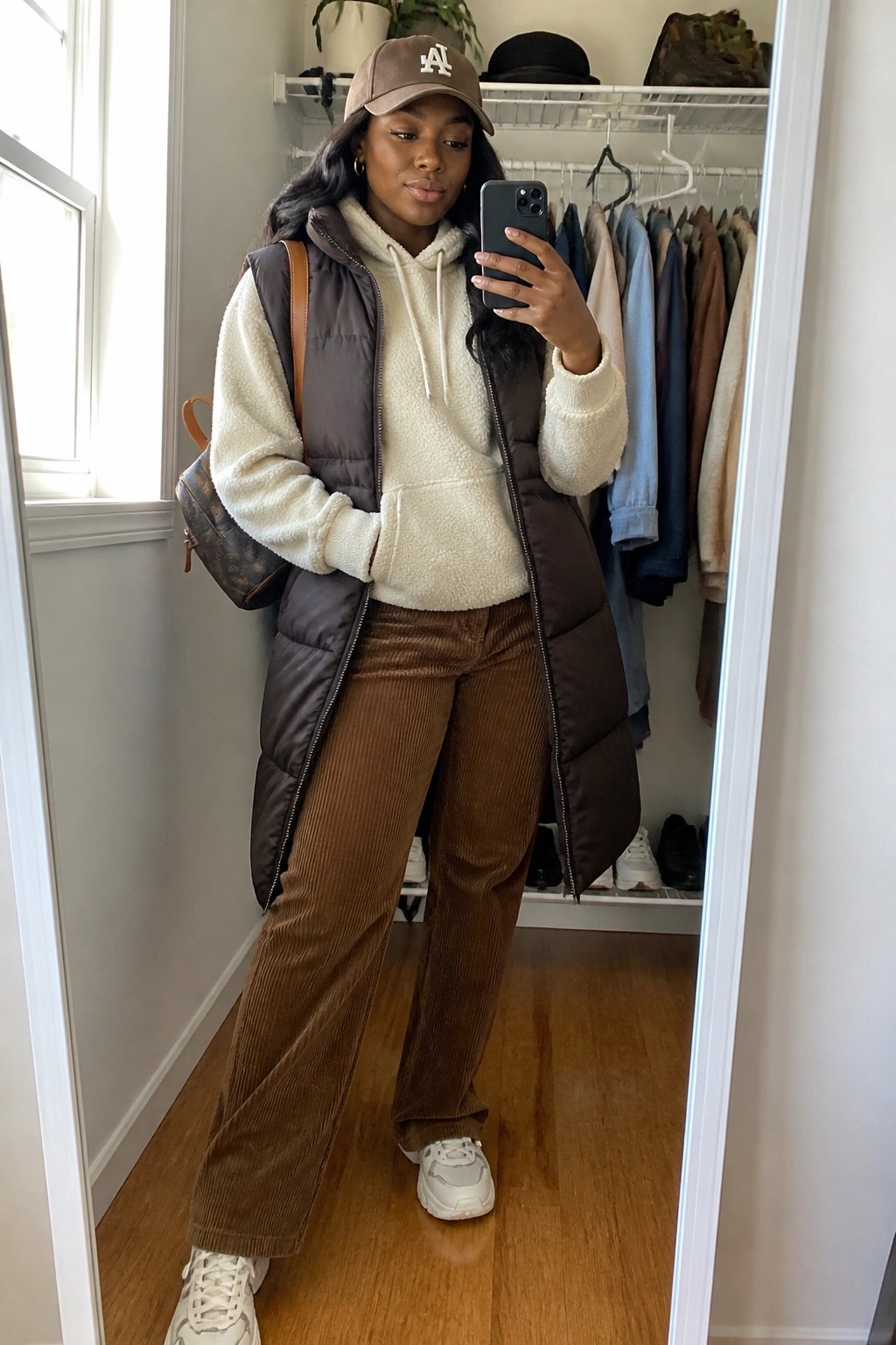 Woman in brown puffer vest, cream hoodie, and corduroy pants taking closet mirror selfie