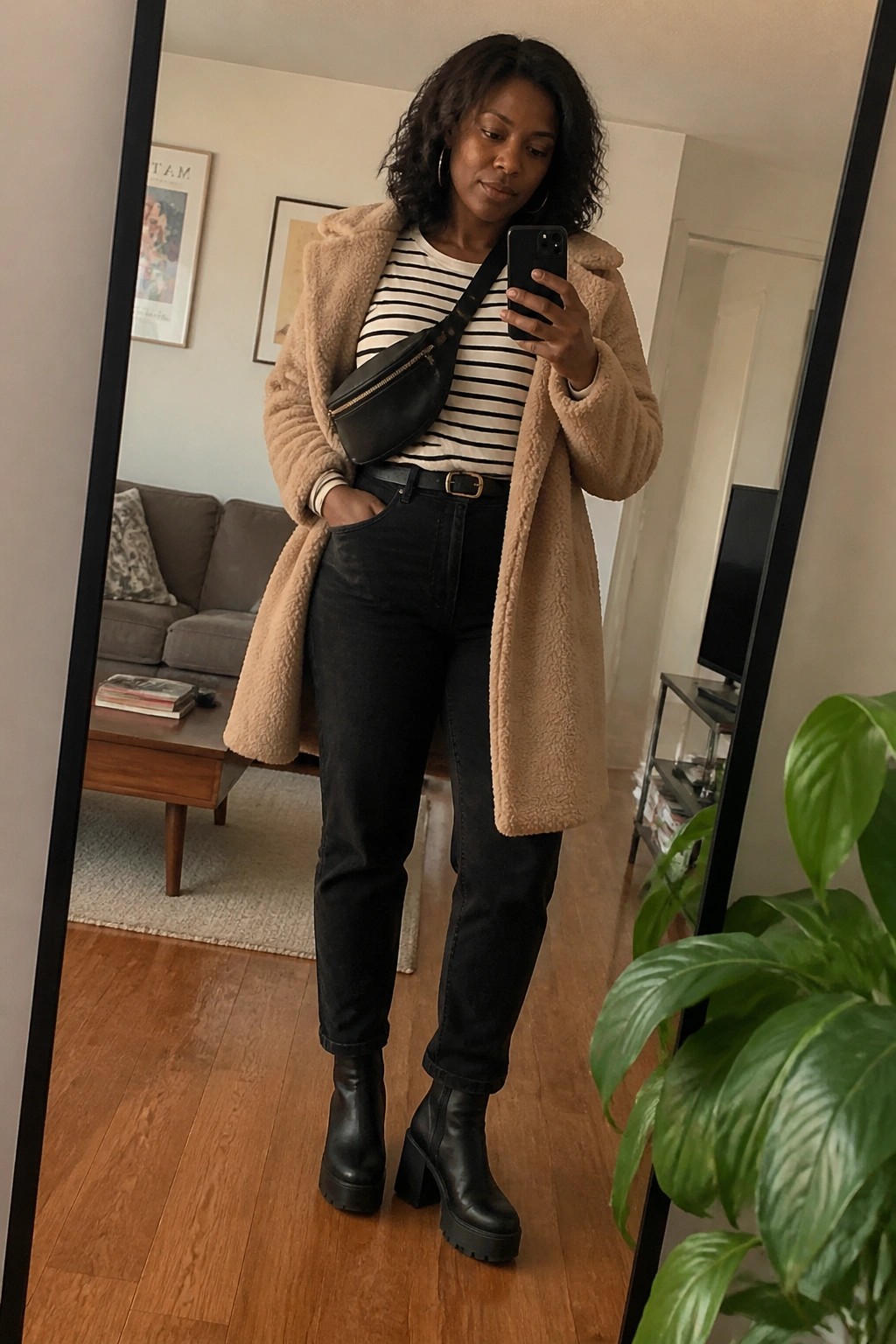 Woman in a beige teddy coat over a black and white striped shirt paired with black jeans a black belt and black platform boots while wearing a black crossbody fanny pack.