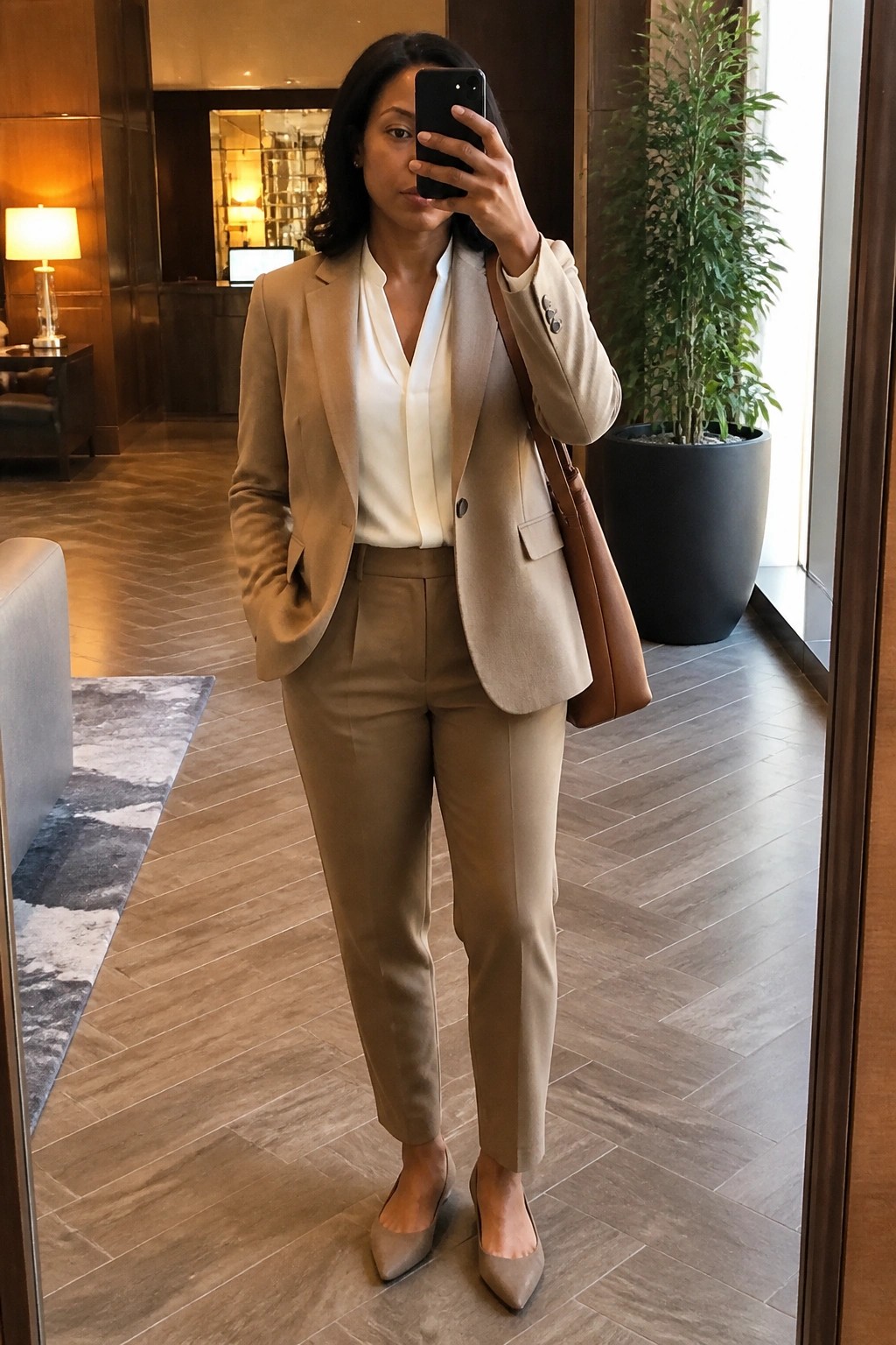 Woman in beige pantsuit taking mirror selfie in modern lobby with handbag