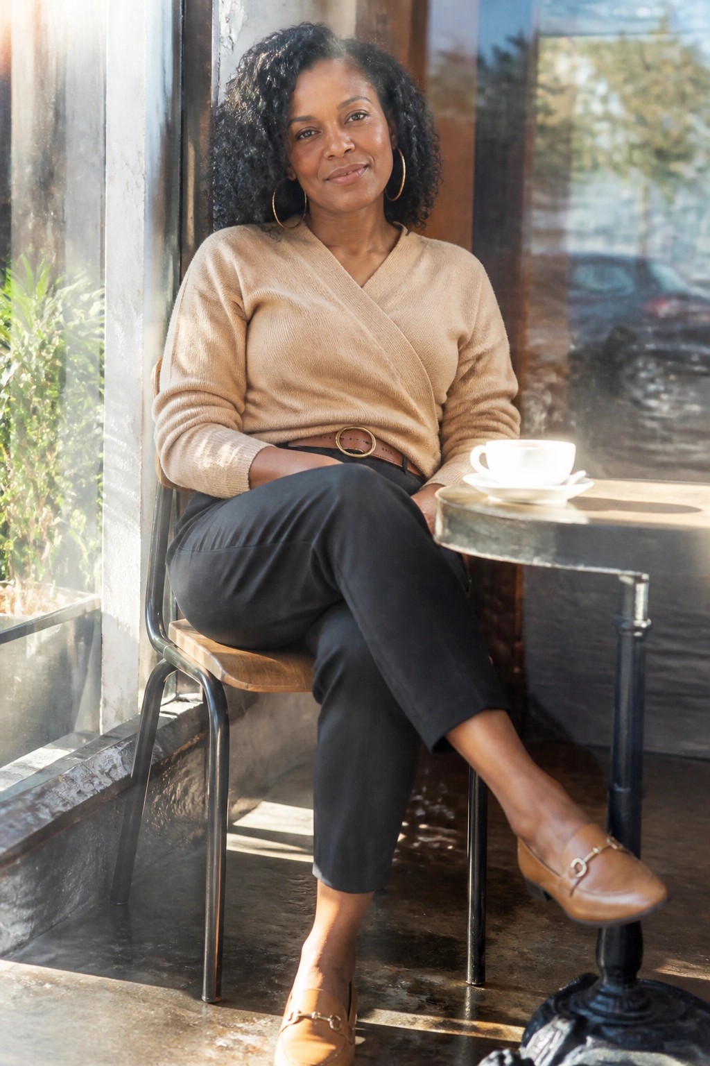 Woman in beige wrap knit sweater, black cropped pants, brown belt with gold buckle, matching brown loafers with gold detail, and large gold hoop earrings.