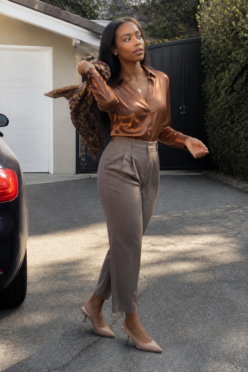 Woman in shiny brown blouse and taupe pants holding scarf by car.