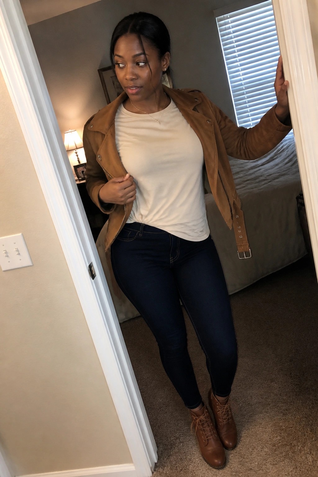 Woman in brown jacket, white tee, and jeans standing in bedroom doorway