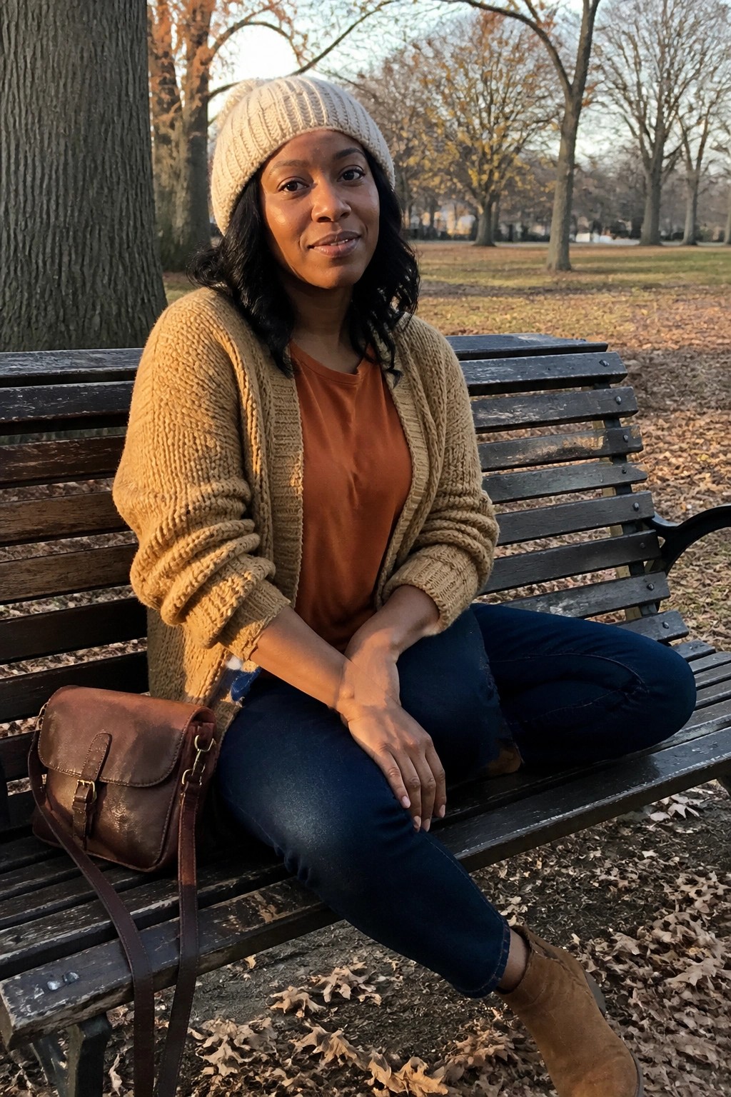 Woman seated on a park bench wearing a beige knit cardigan over a rust orange tee, dark blue jeans, brown suede boots, and a matching beige beanie with a brown leather crossbody bag.
