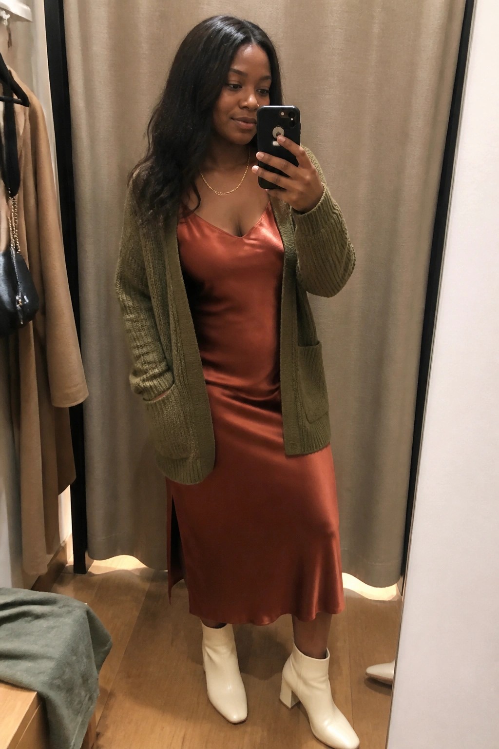 Woman in a mirror selfie wearing an olive green chunky knit cardigan over a rust orange satin midi slip dress with white ankle boots and a thin gold necklace.