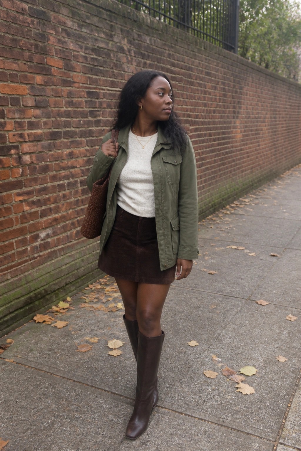 Woman in green jacket and brown corduroy skirt by brick wall with autumn leaves
