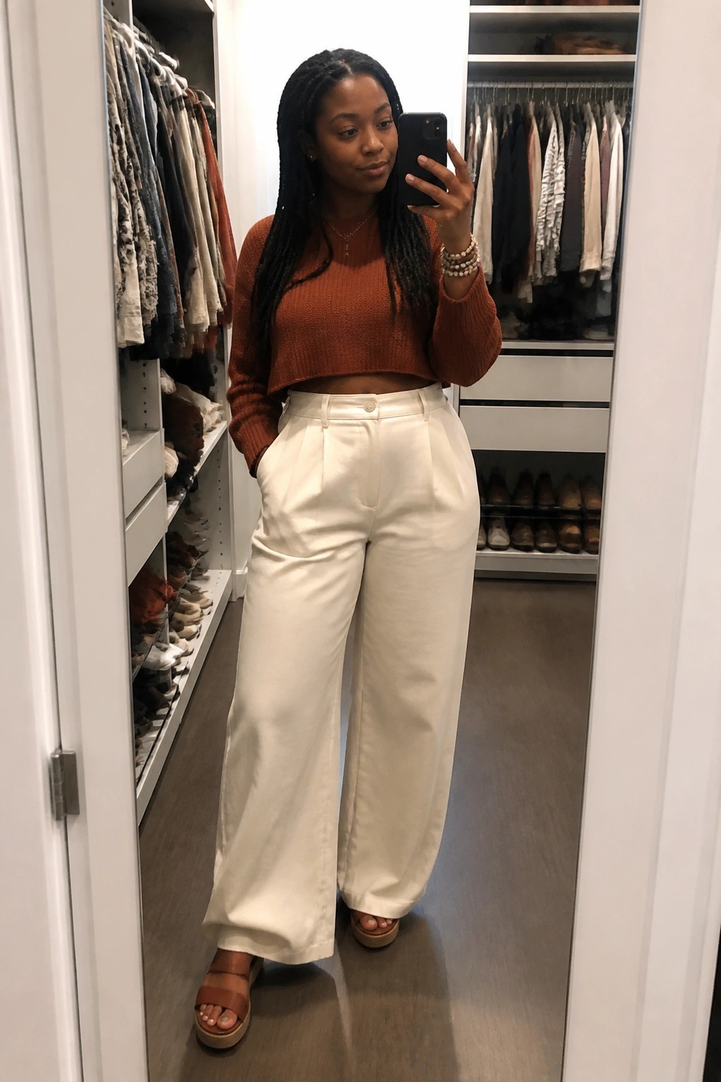 Woman with braids in rust crop sweater and white wide-leg pants taking closet mirror selfie
