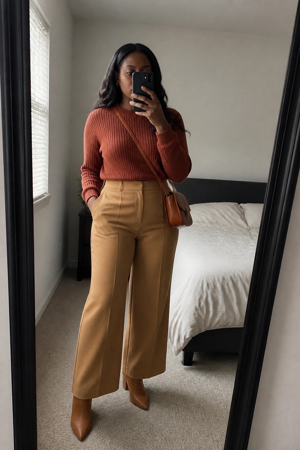 Woman in rust sweater and tan wide-leg pants taking bedroom mirror selfie