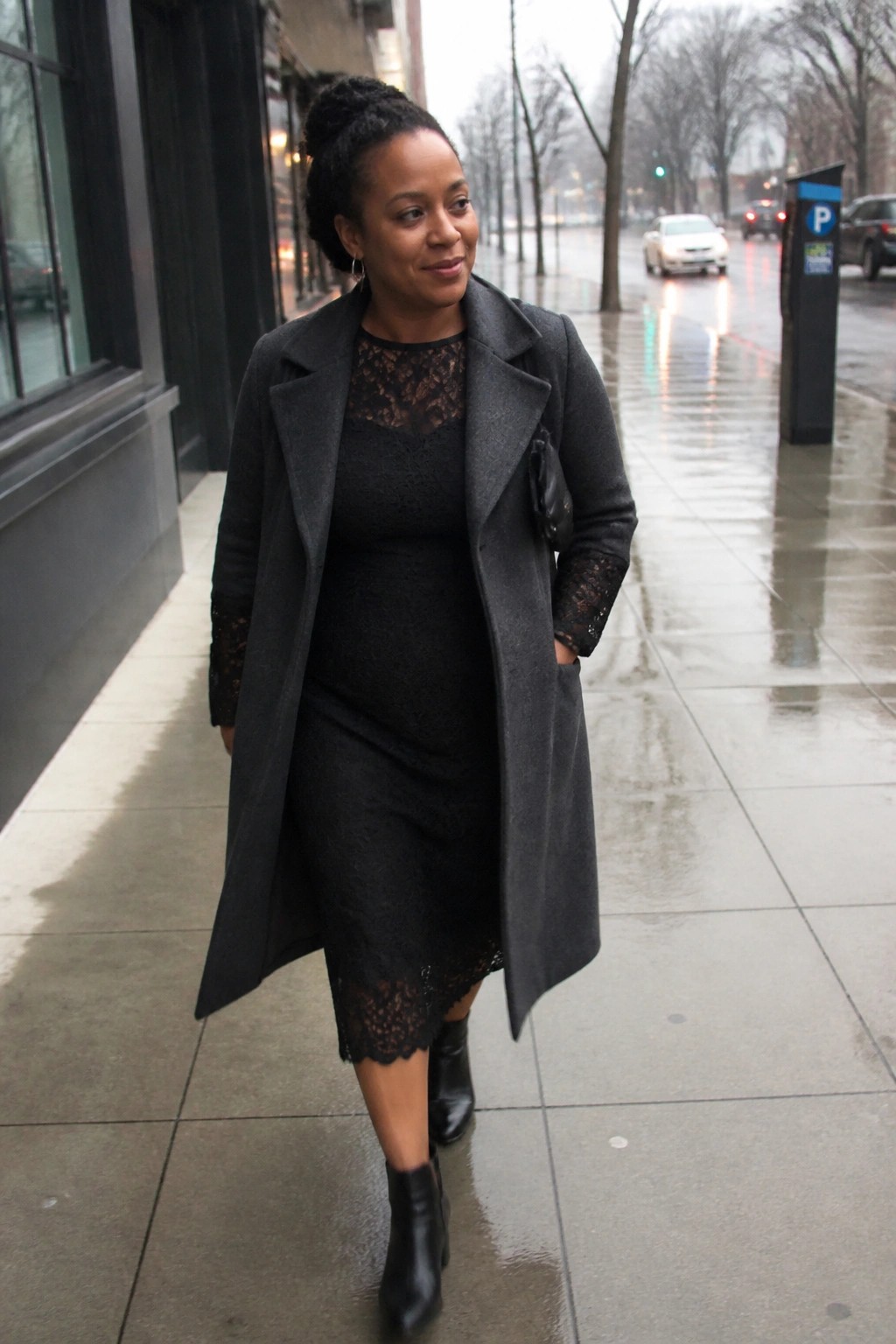 Woman in black lace dress and gray coat walking on wet sidewalk.