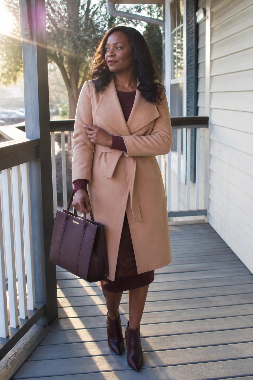 Woman in tan belted coat and burgundy dress with handbag on porch