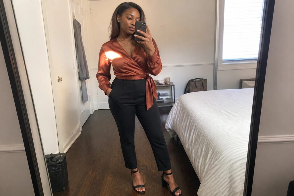 Woman in rust satin wrap blouse tied at waist paired with black straight leg trousers and black heeled sandals taking a mirror selfie.