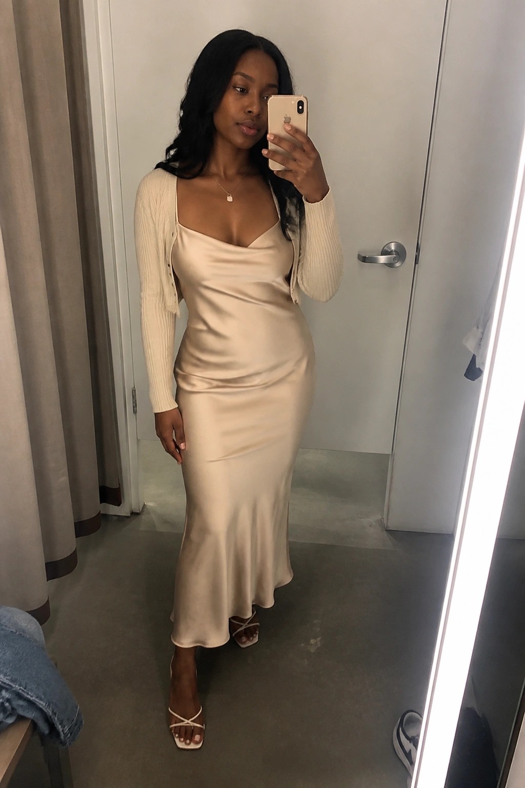 Woman in beige satin slip dress and cardigan taking mirror selfie with iPhone