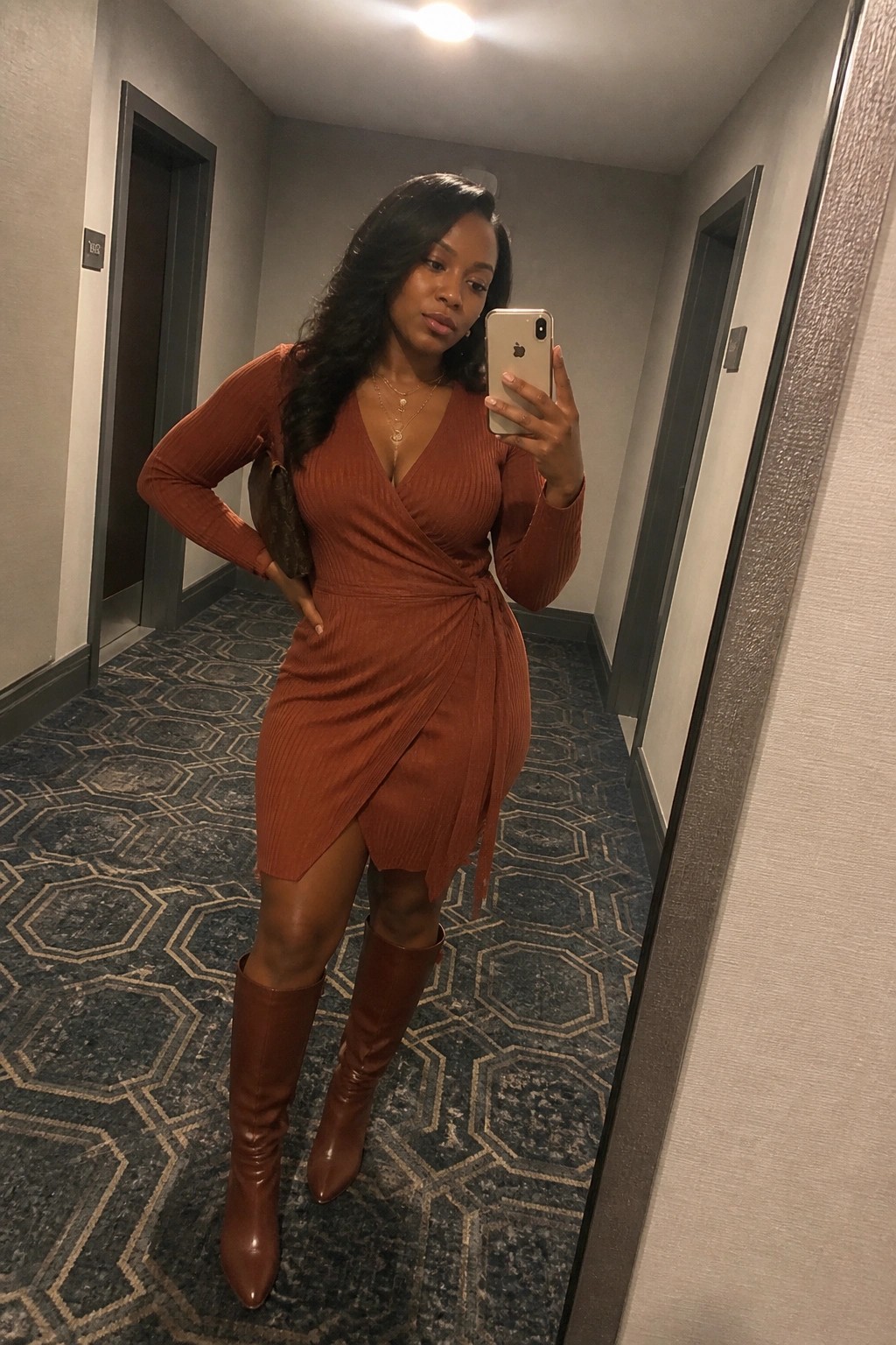 Woman in rust wrap dress and brown boots taking mirror selfie in hallway