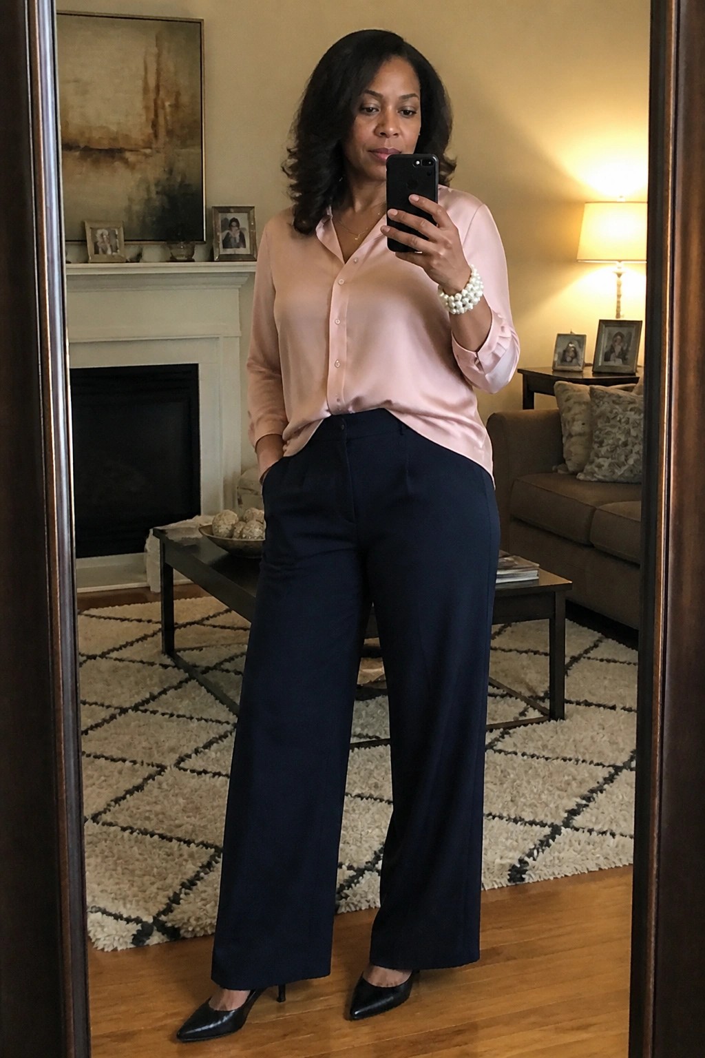 Woman wearing a light pink satin button down blouse tucked into navy wide leg trousers with black pointed heels and a pearl bracelet while taking a mirror selfie.