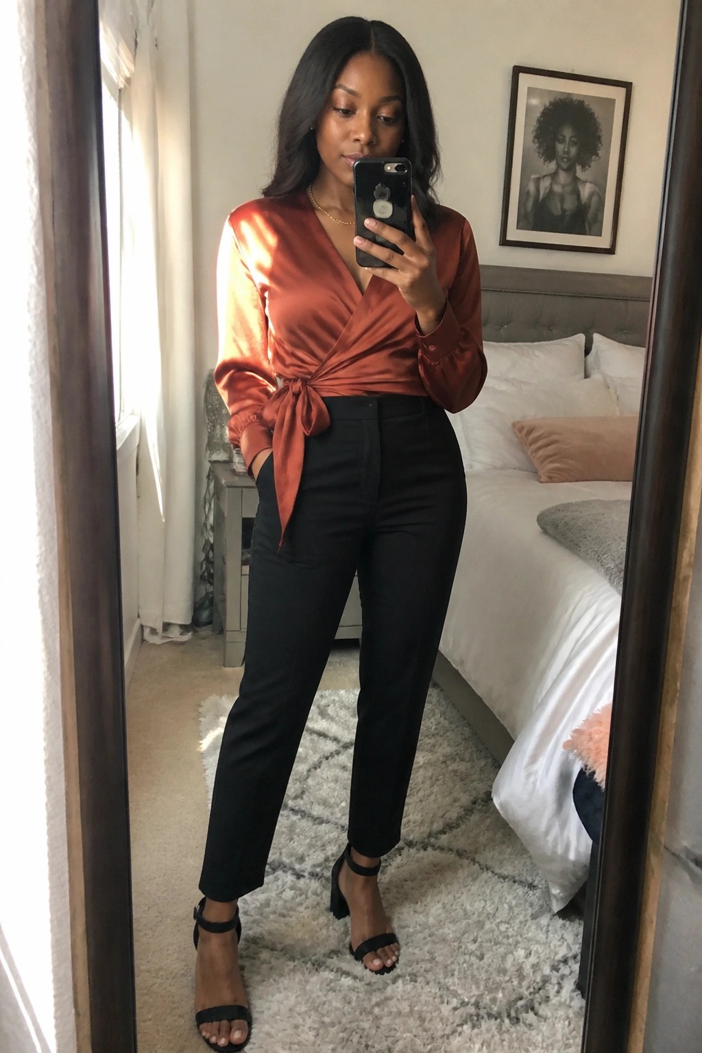 Woman in rust satin wrap blouse tied at waist paired with black straight leg trousers and black heeled sandals taking a mirror selfie.