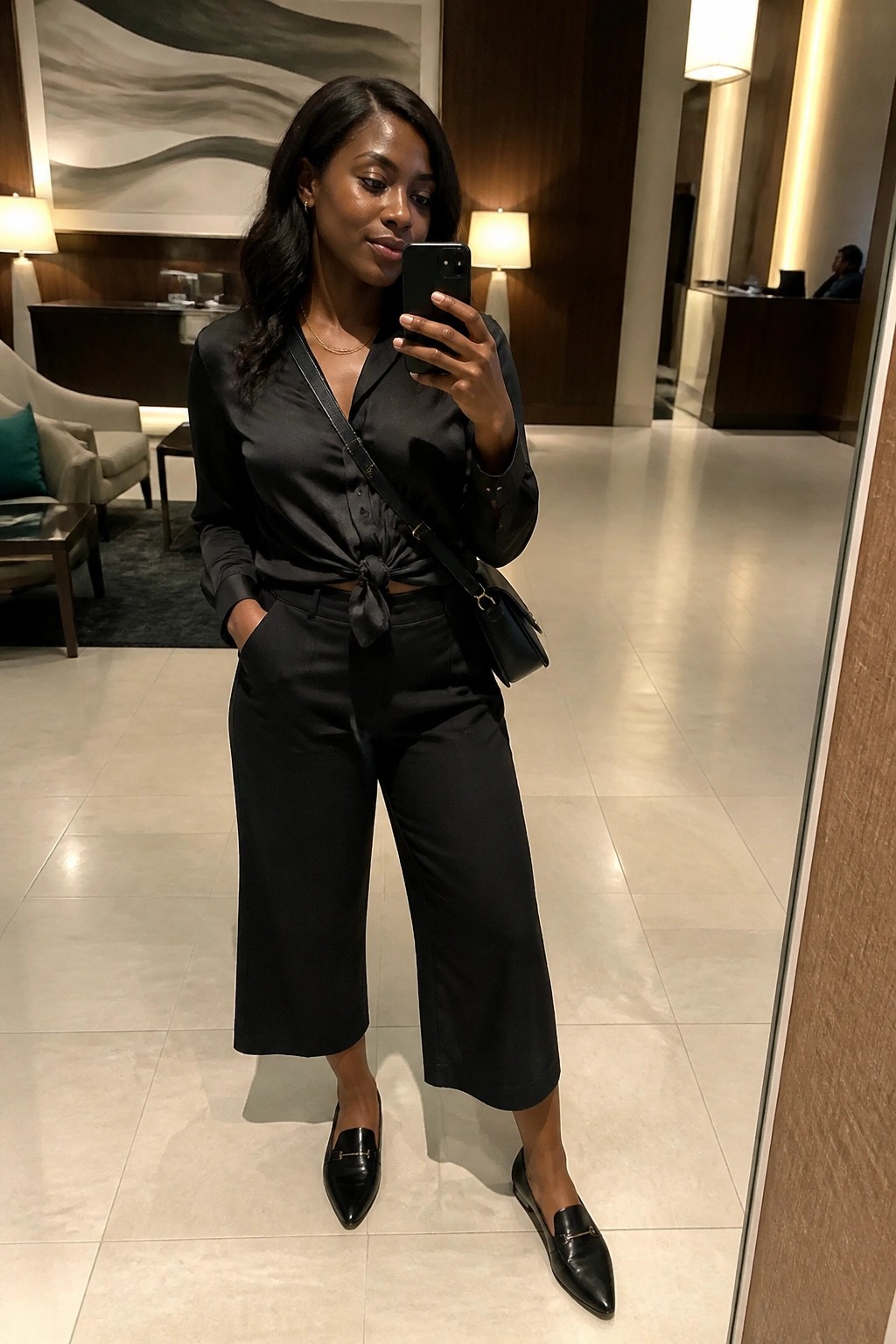 Woman in black satin tied blouse and cropped pants taking mirror selfie in lobby