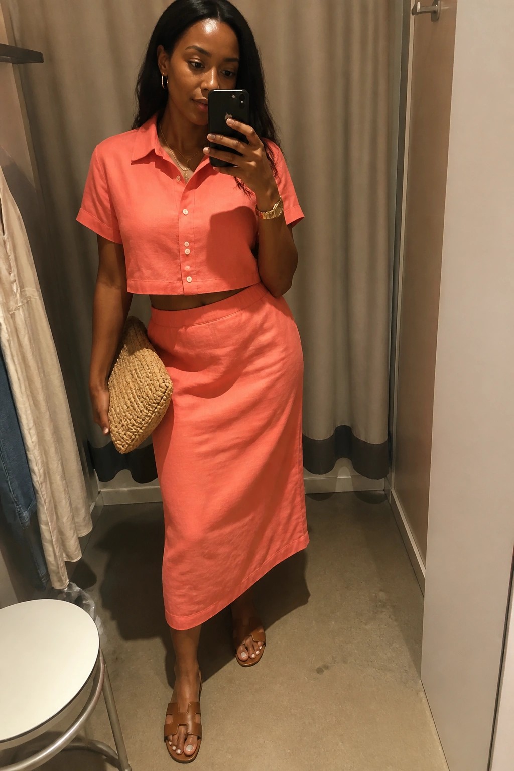 Woman wearing a matching coral linen cropped button-up shirt and midi skirt with brown leather sandals, a woven clutch, and gold jewelry in a fitting room mirror selfie.