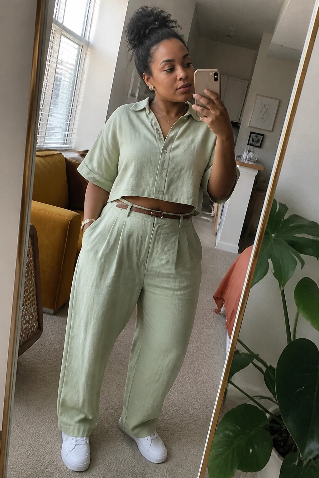 Woman in light green cropped shirt and pleated pants taking mirror selfie.