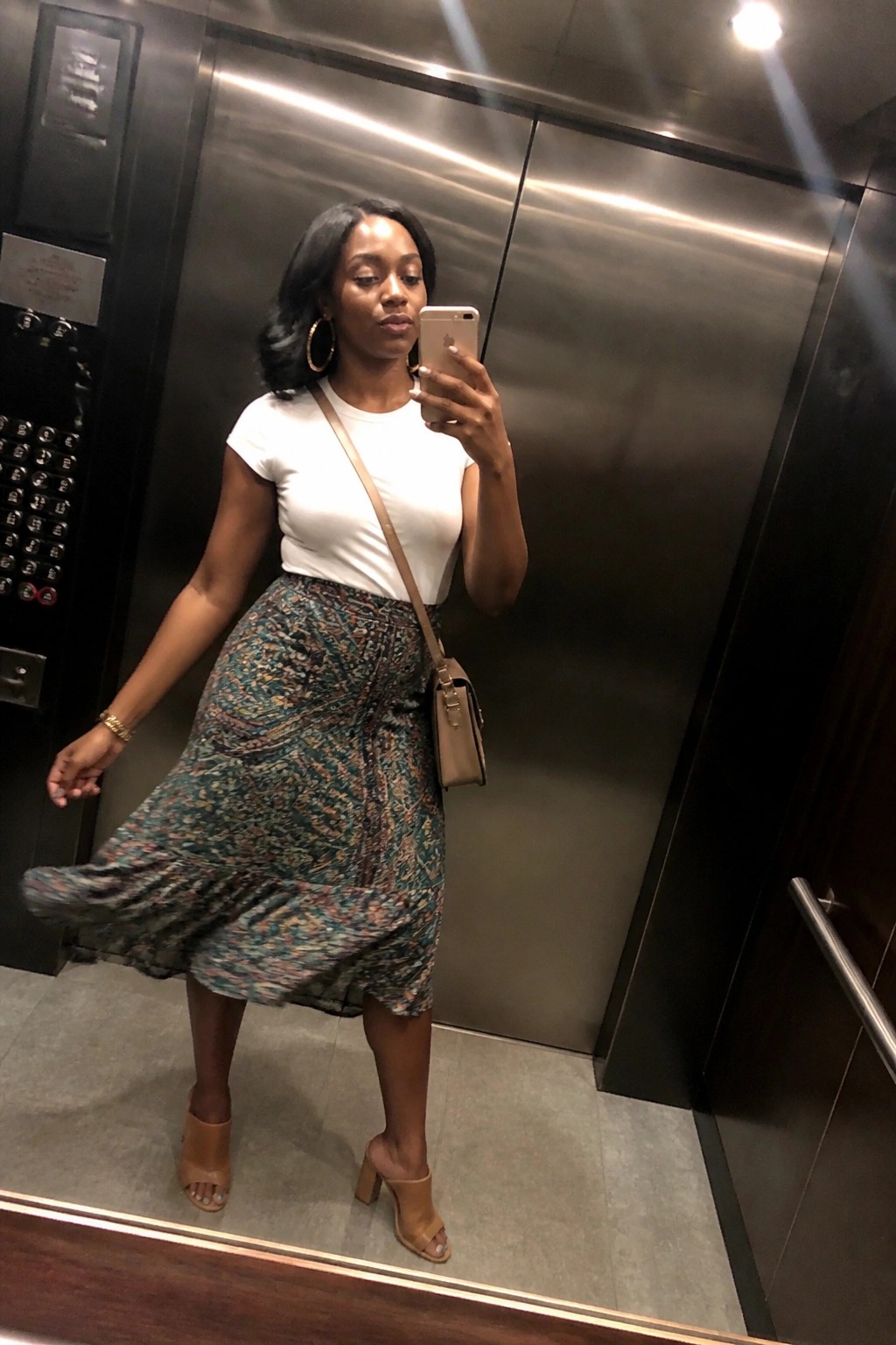 Woman in white top and patterned skirt taking mirror selfie in elevator