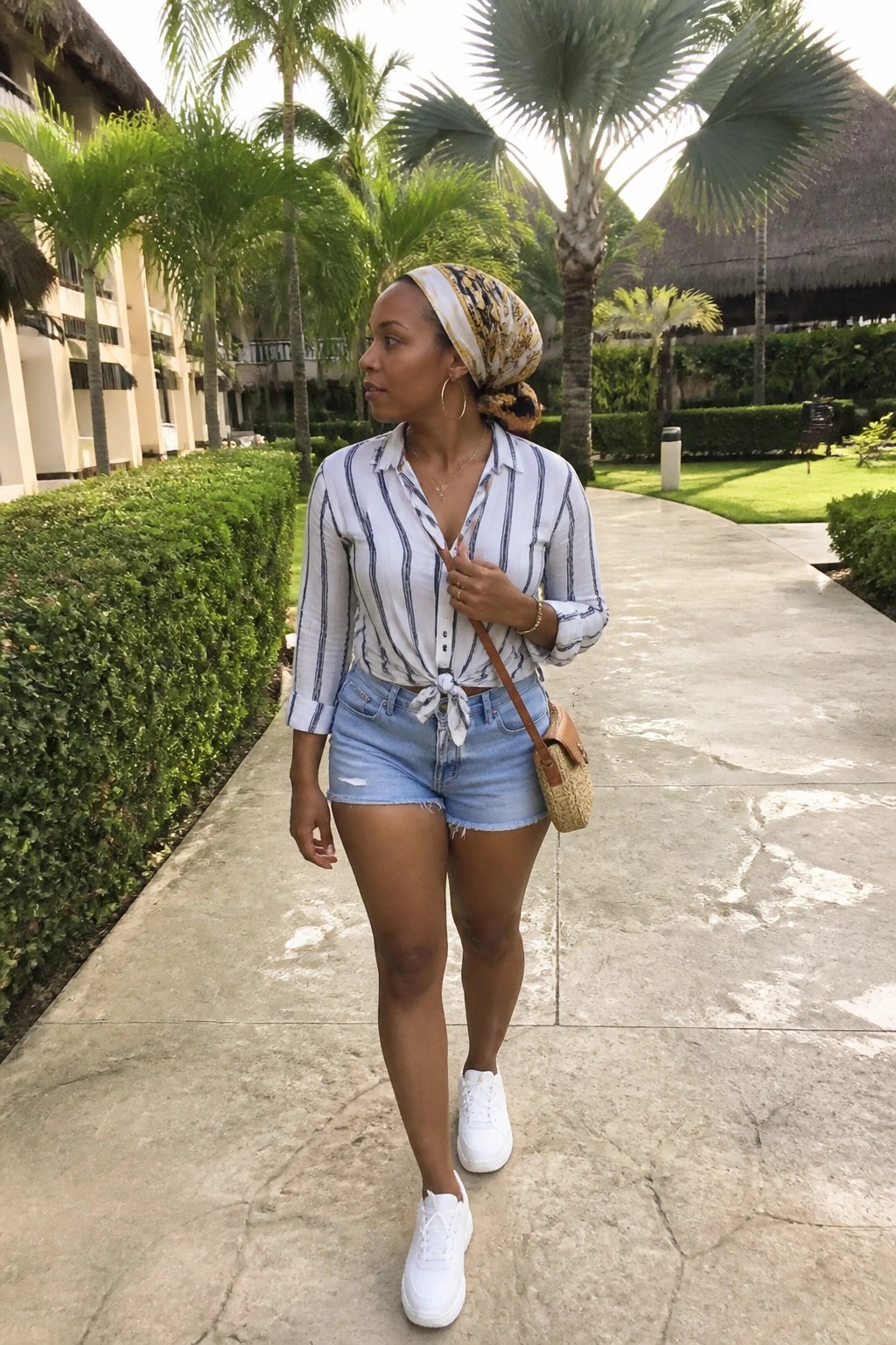 Woman in a white and navy striped button down shirt tied at the waist, light blue denim shorts, white sneakers, patterned headscarf, and woven crossbody bag standing on a paved path.