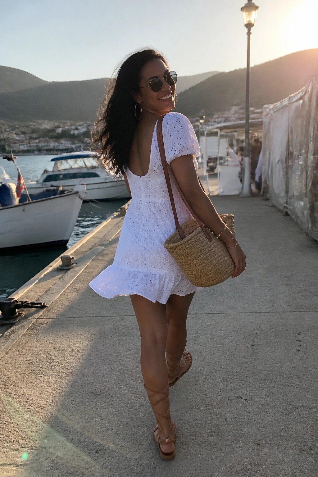 Woman in white lace dress with straw bag on harbor pier at sunset.
