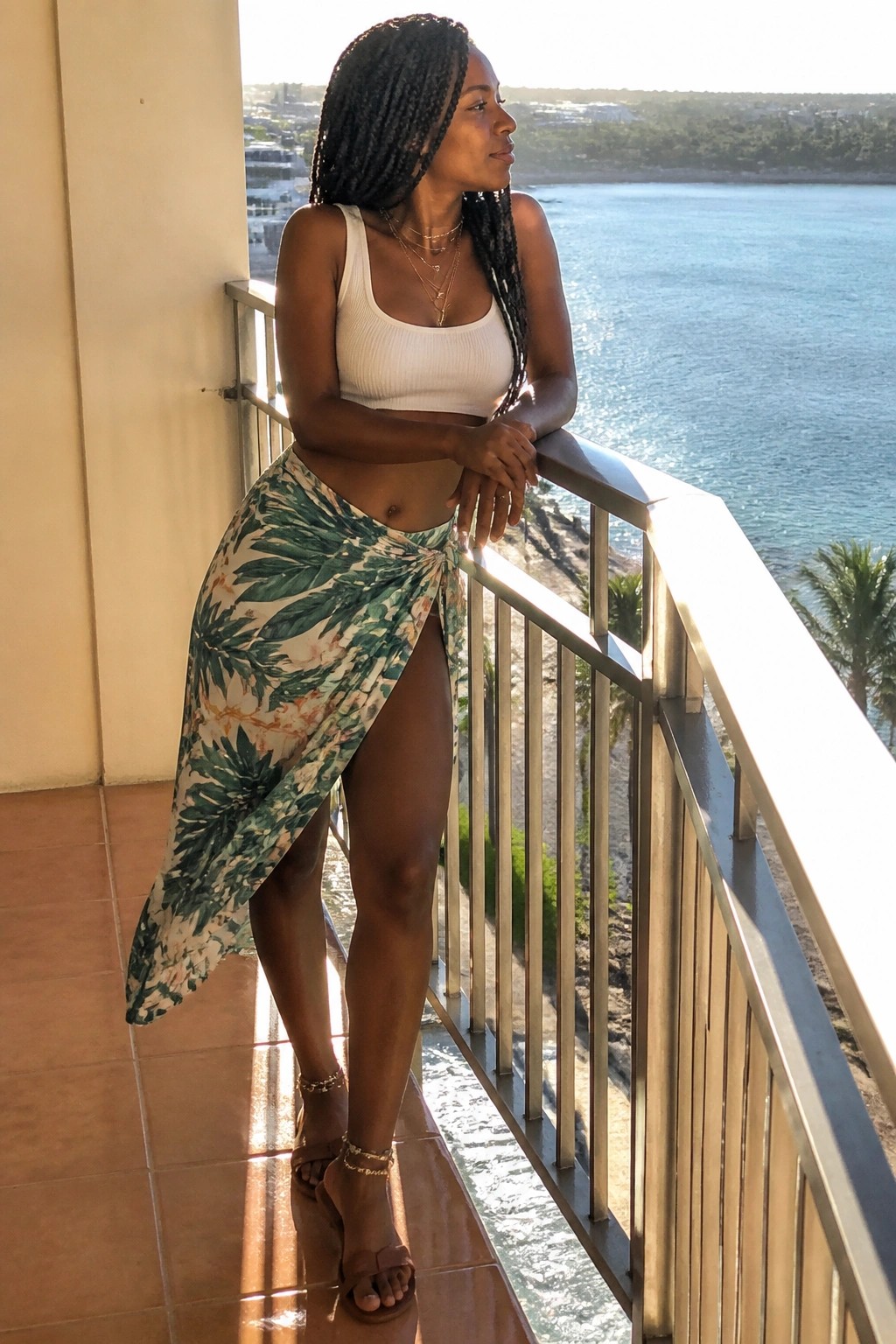 Black woman wearing a white ribbed crop tank top with a green and white tropical leaf print wrap skirt featuring a high side slit, layered gold necklaces, gold anklets, and brown flat sandals on a tiled balcony.