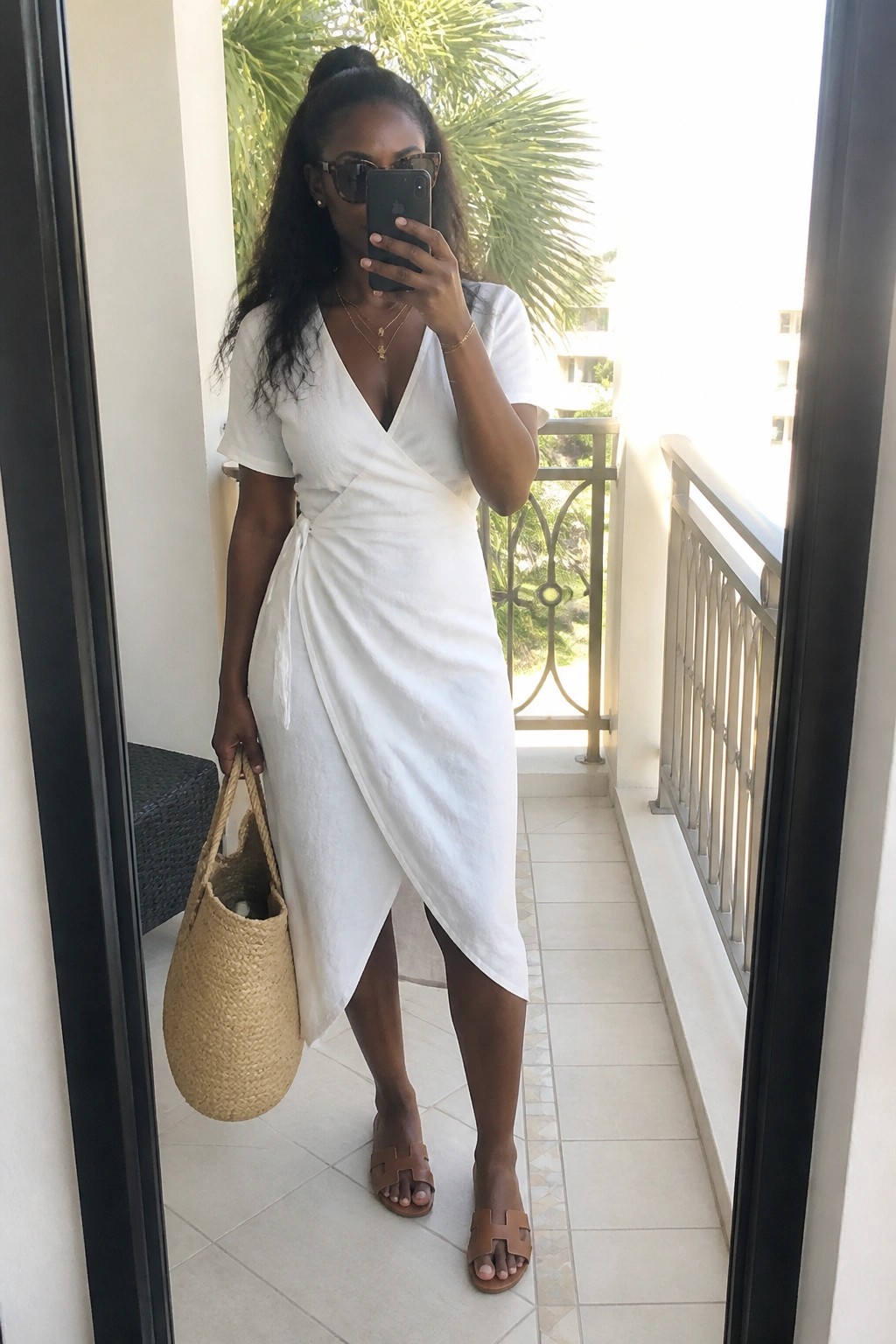 Black woman wearing a white short sleeve wrap dress with asymmetrical hem, brown leather slide sandals, gold layered necklaces, sunglasses, and holding a woven straw tote bag while taking a mirror selfie on a balcony.