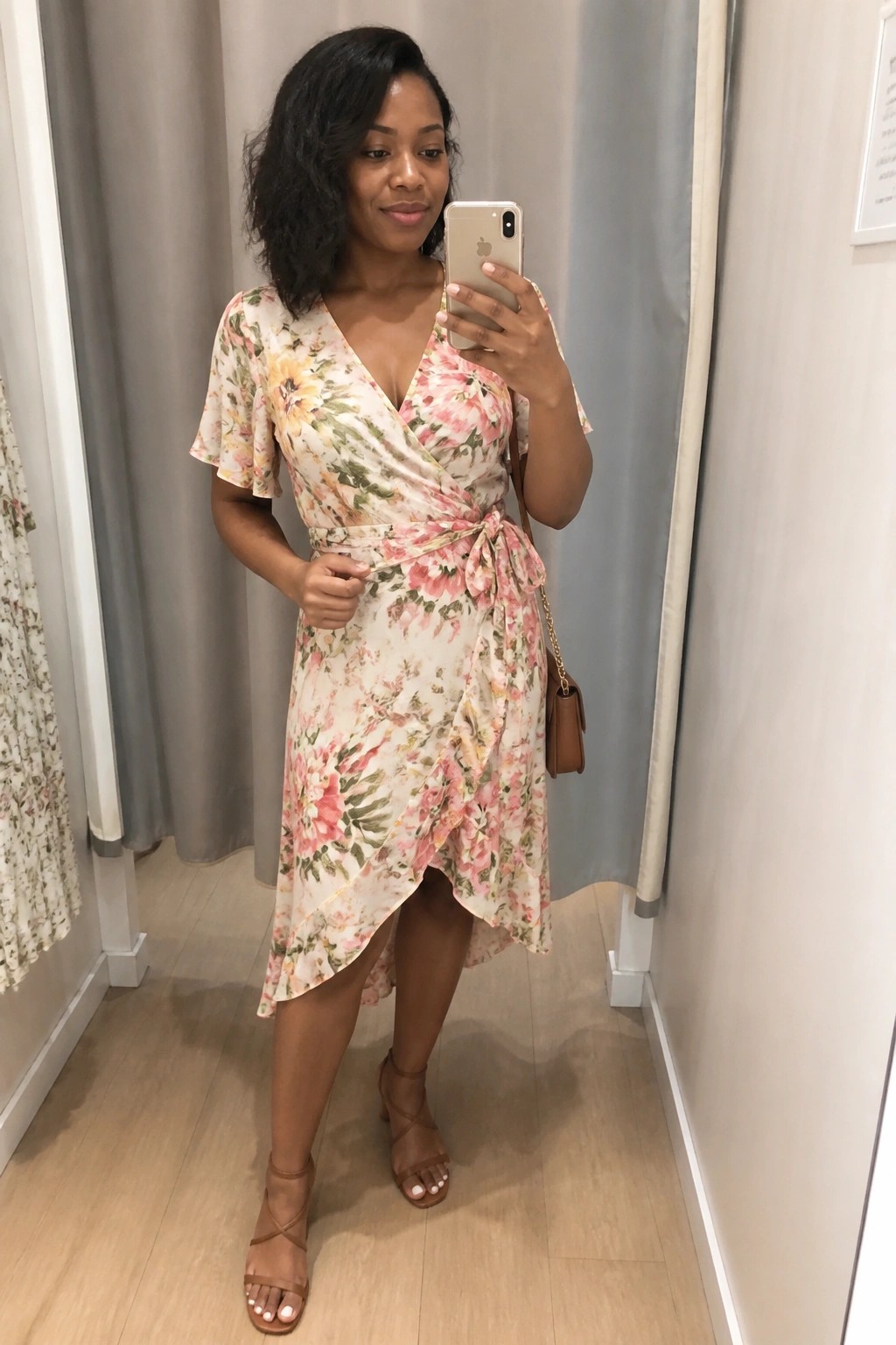 Woman wearing a light floral wrap dress with short sleeves and a tied waist paired with brown strappy sandals and a small brown crossbody bag while taking a mirror selfie.