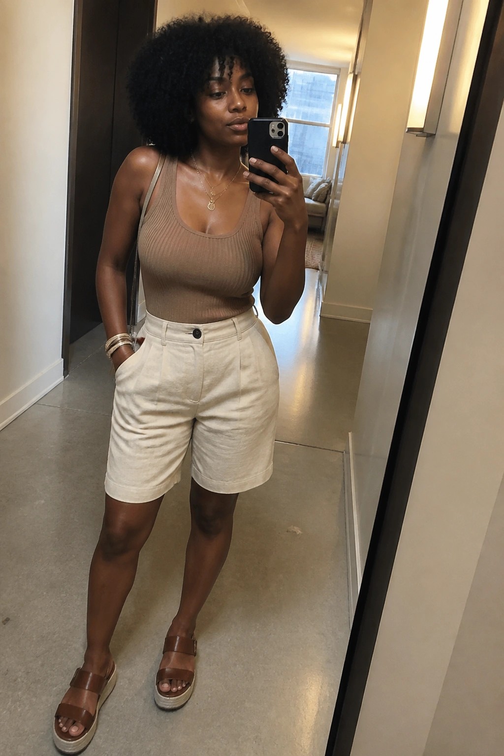 Woman in beige tank top and white shorts taking mirror selfie in hallway