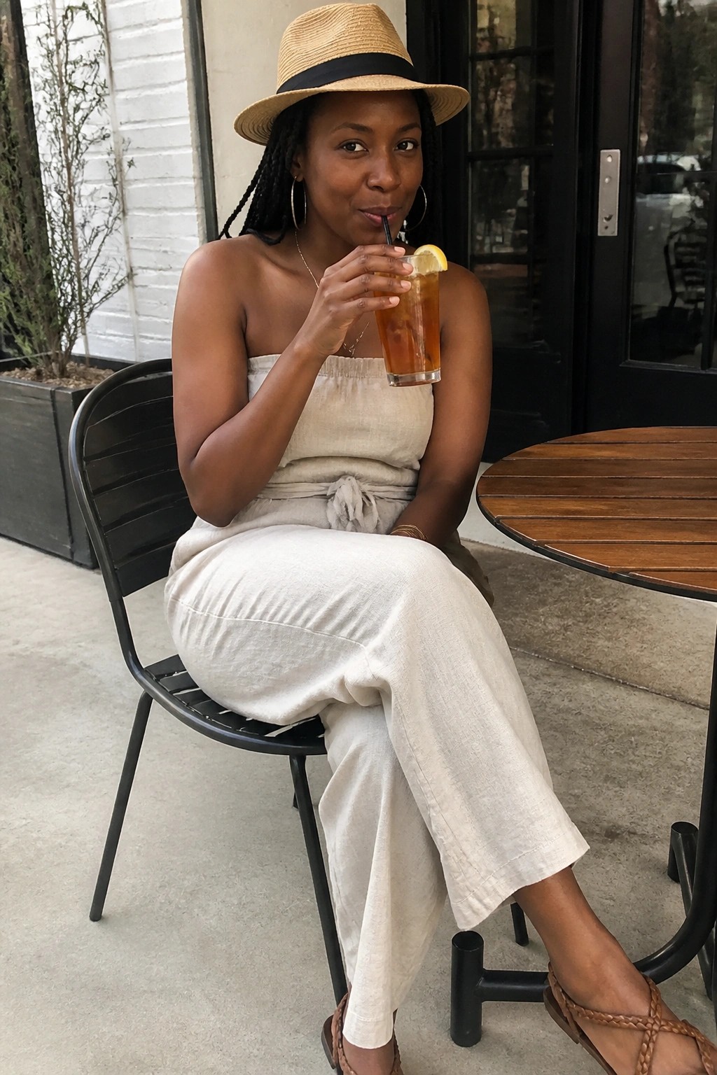 Woman in beige strapless wide-leg jumpsuit with waist tie, straw hat, hoop earrings, and brown sandals, seated at outdoor table holding iced drink.
