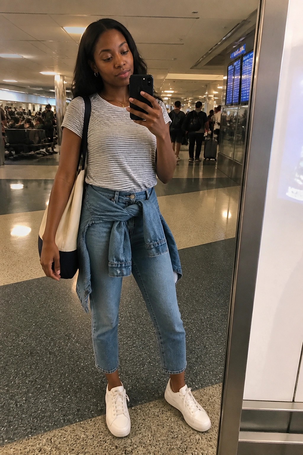 Woman in striped shirt and jeans taking mirror selfie in airport terminal