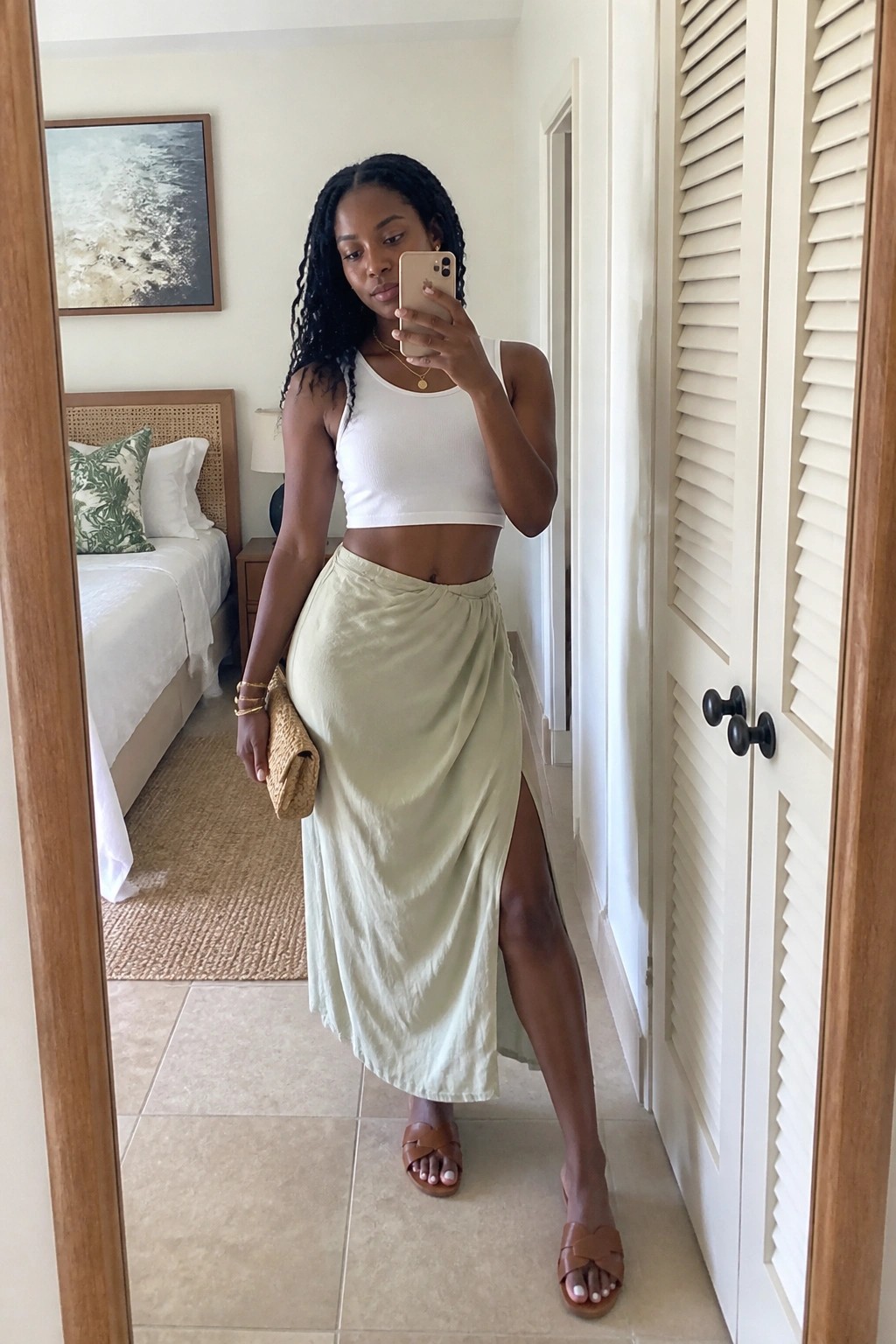 Black woman in mirror selfie wearing white ribbed crop top, pale green satin wrap skirt with side slit, brown slide sandals, and gold jewelry while holding a woven clutch