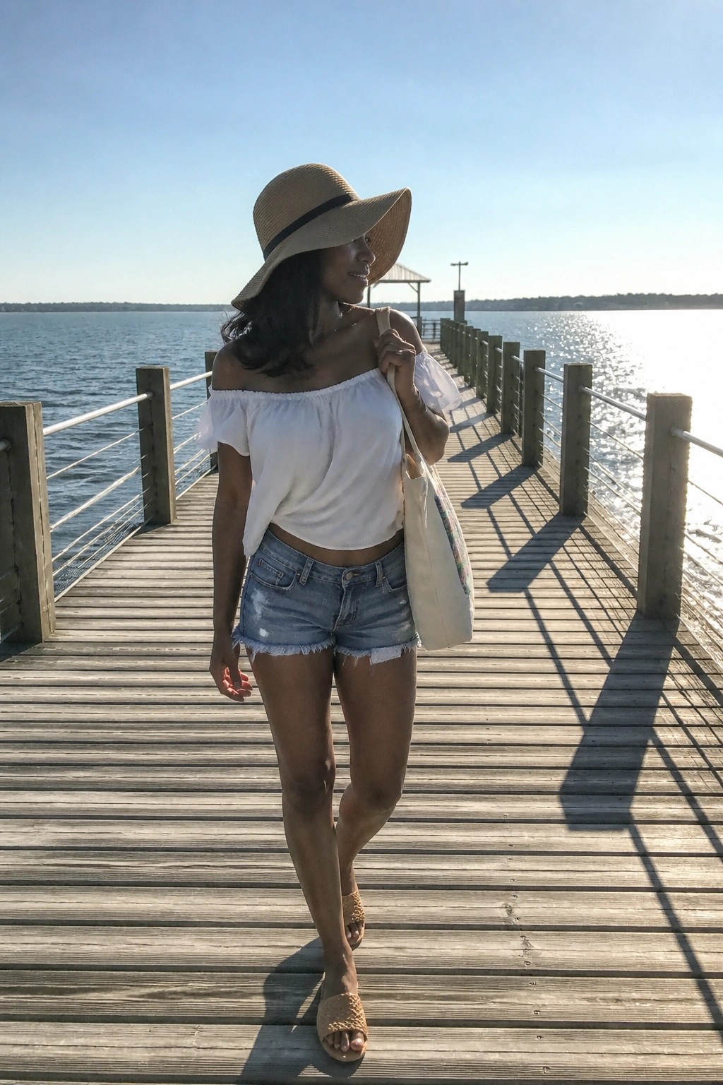 Woman in white off-shoulder ruffled top, light-wash distressed denim shorts, straw wide-brim hat, beige tote bag, and woven slide sandals standing on wooden pier.