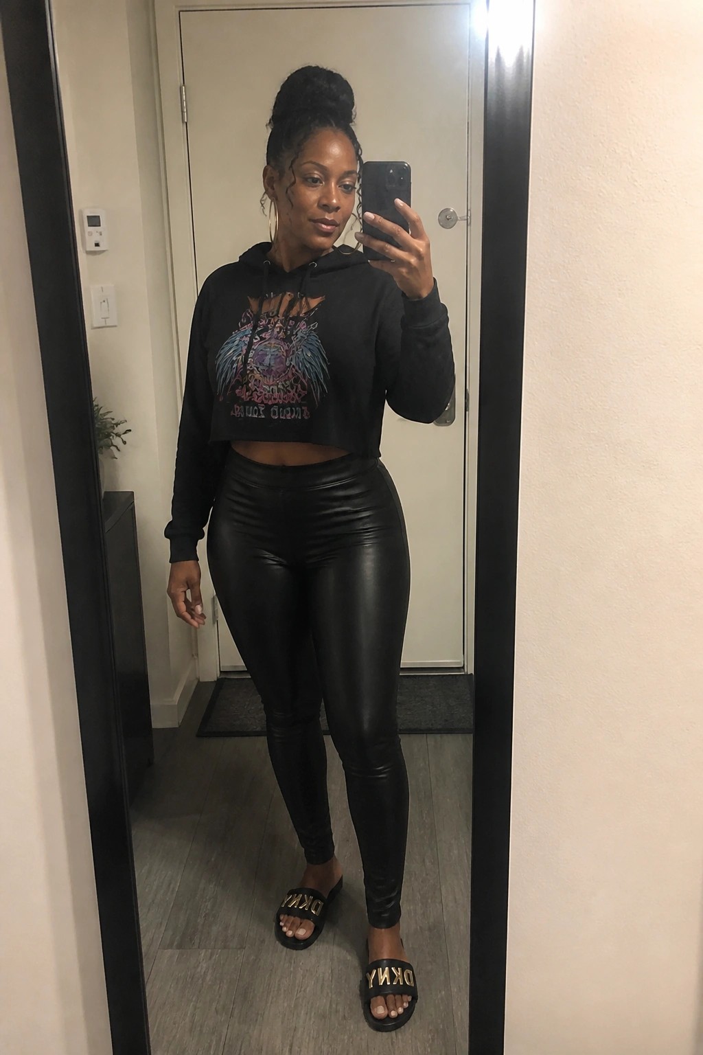 Woman in black cropped graphic hoodie, shiny black leggings, and black DKNY slides taking a mirror selfie in an indoor hallway.