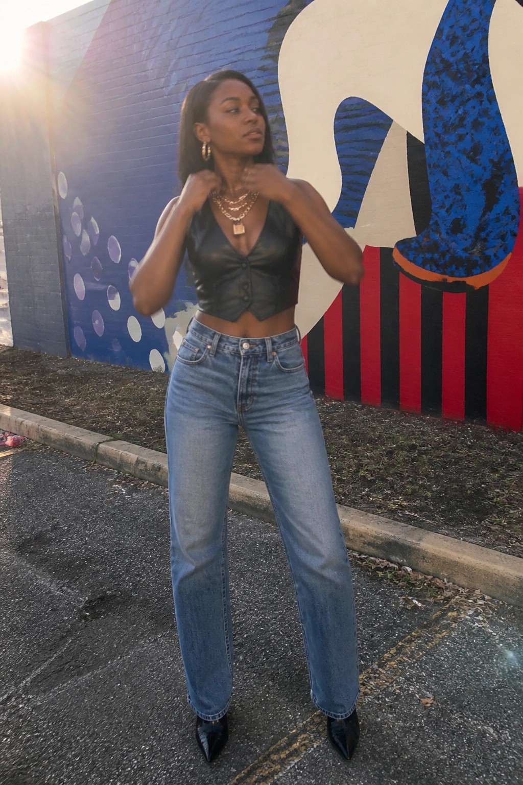Woman in black crop top and blue jeans adjusts necklaces before colorful mural.